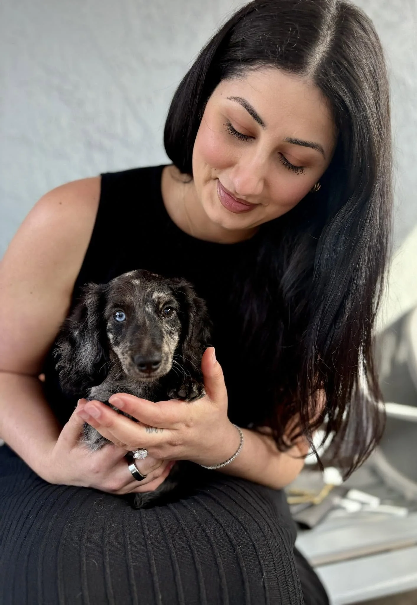 A woman with long black hair holding a black and gray puppy with bright blue eyes.