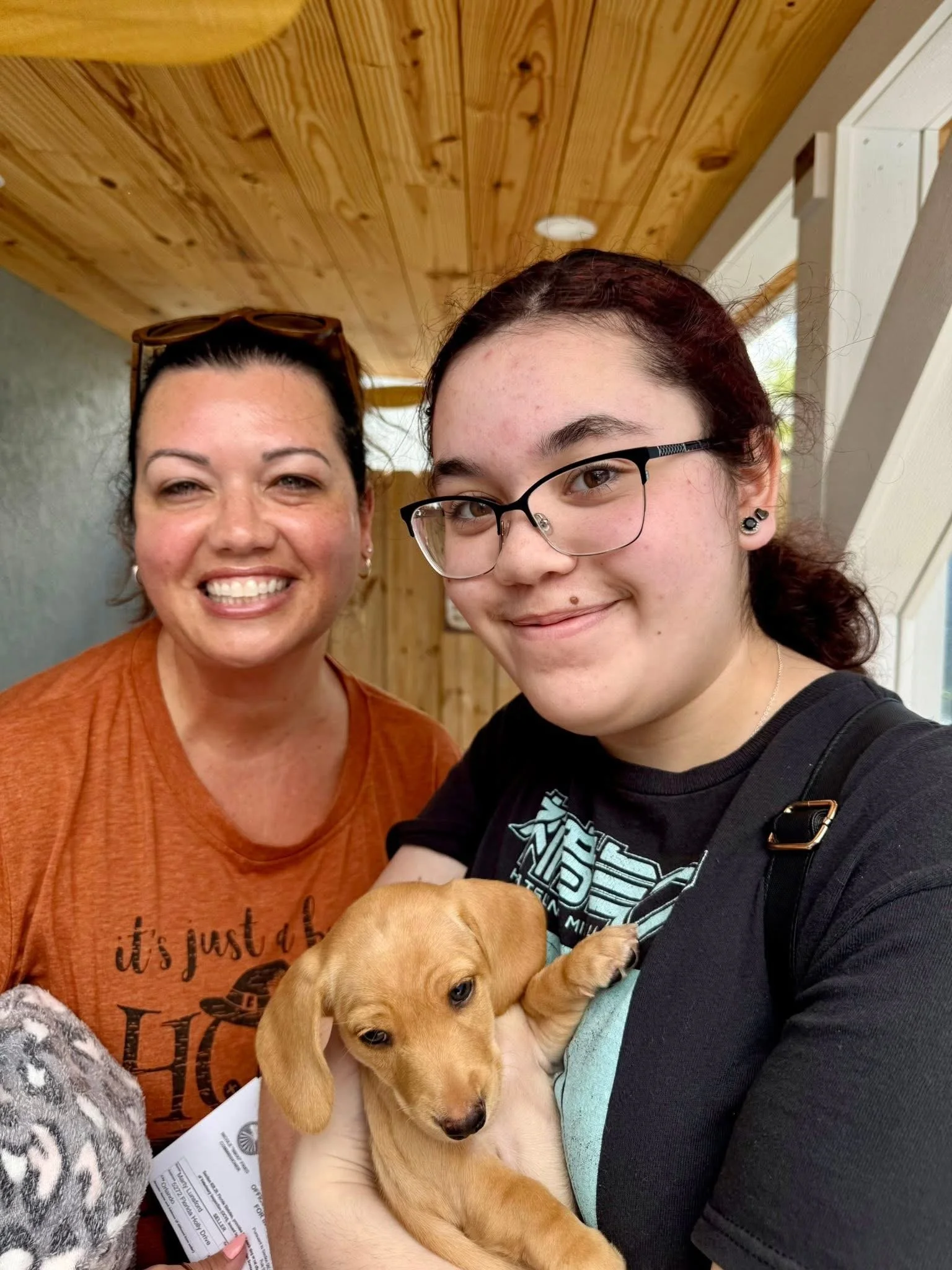 Two women are smiling and holding a small tan puppy. One woman has glasses and dark hair, the other is older with dark hair and sunglasses on her head. They are indoors with a wooden ceiling.