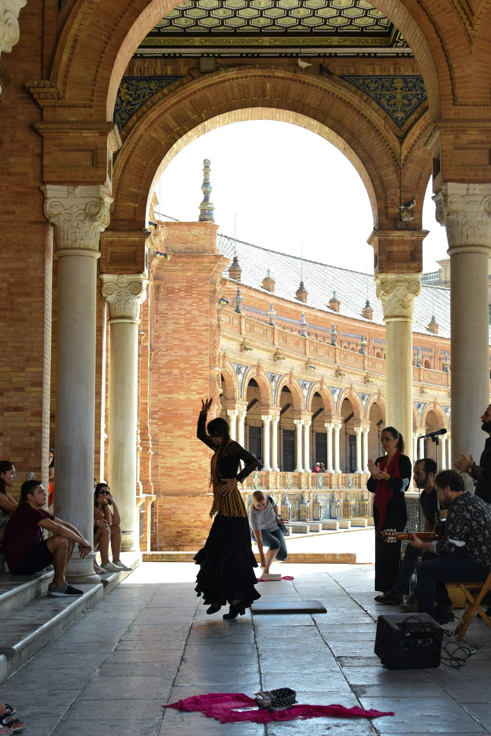 Flamenco dancers performing in an outdoor courtyard with audience seated along columns. The courtyard has brick walls, arches, and a decorative ceiling, with a person playing guitar and another clapping.