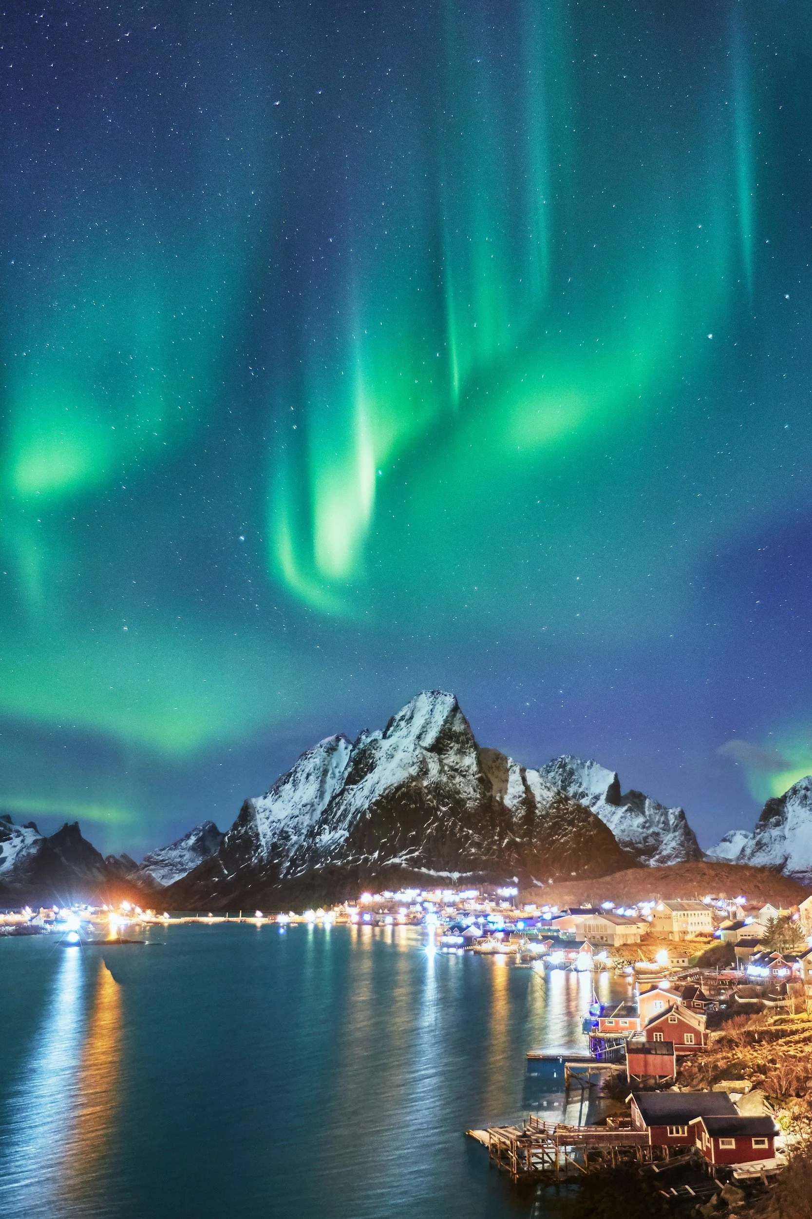Nighttime view of a snowy mountain range with the Northern Lights in the sky. There is a small town by the water, with houses and boats, illuminated with bright lights, reflecting on the water.