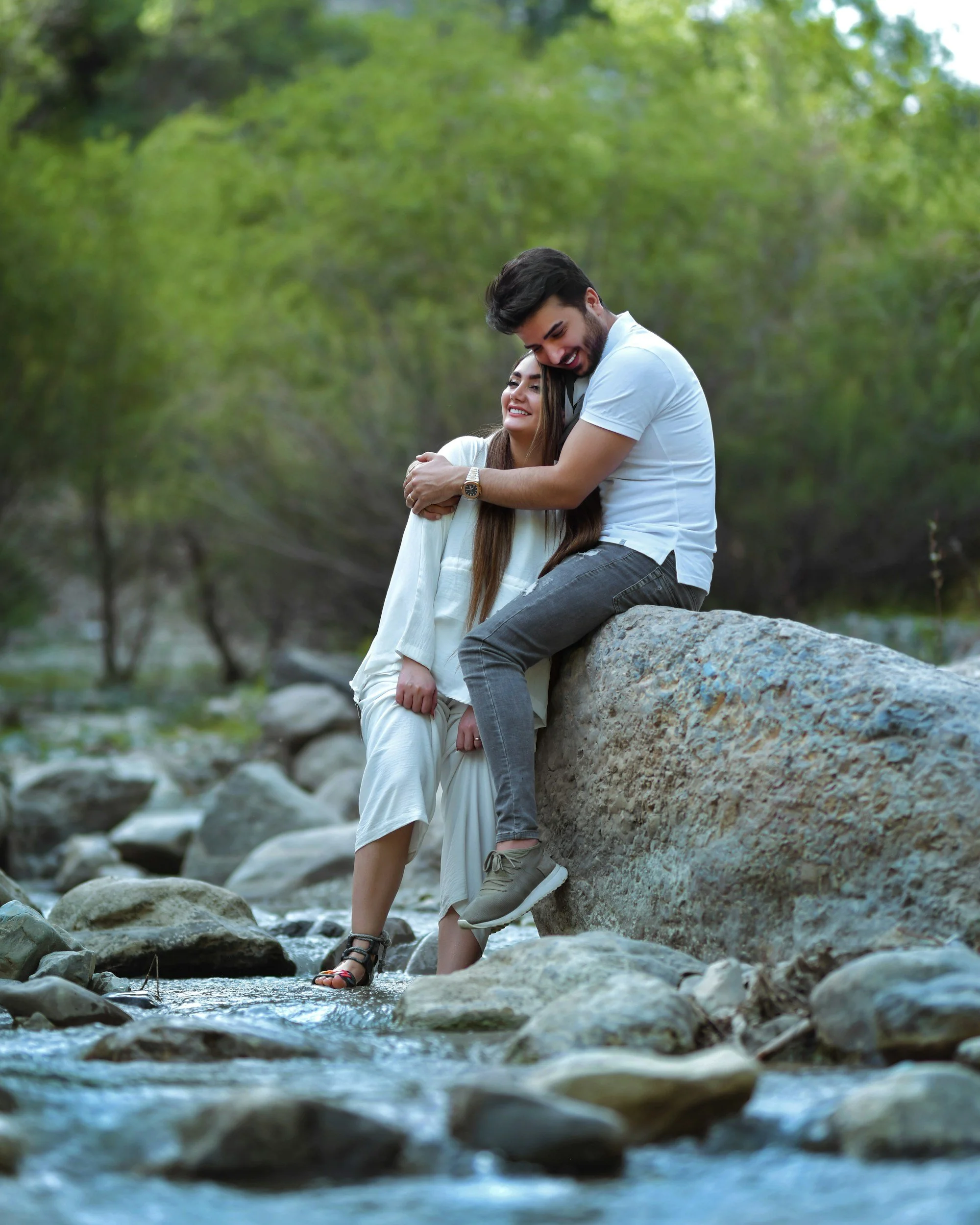 A happy couple hugging on a large rock by a creek in a wooded area.