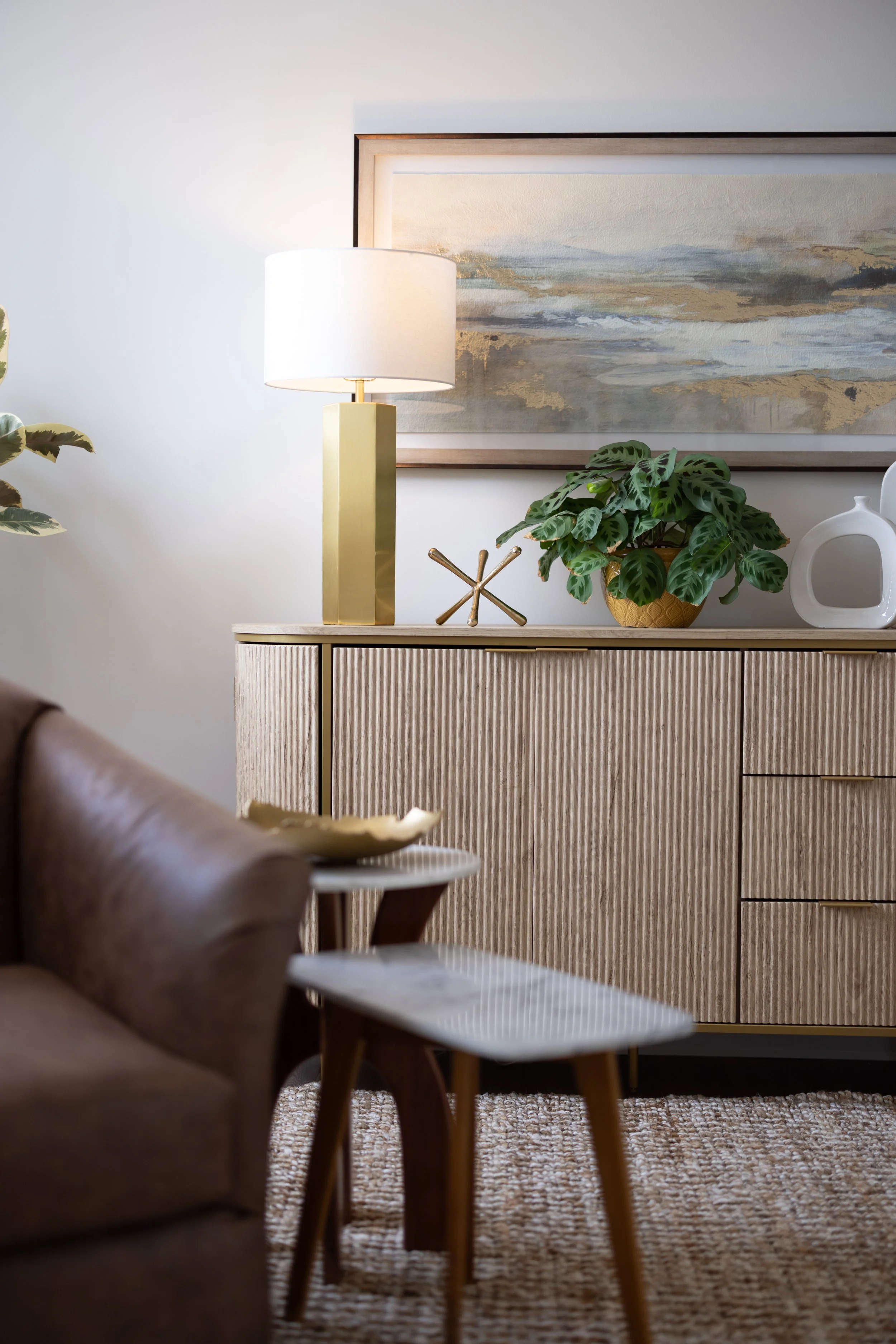 Living room with a sideboard, plants, a painting, and a lamp on top; blurred armchair and side table with a bowl in the foreground.
