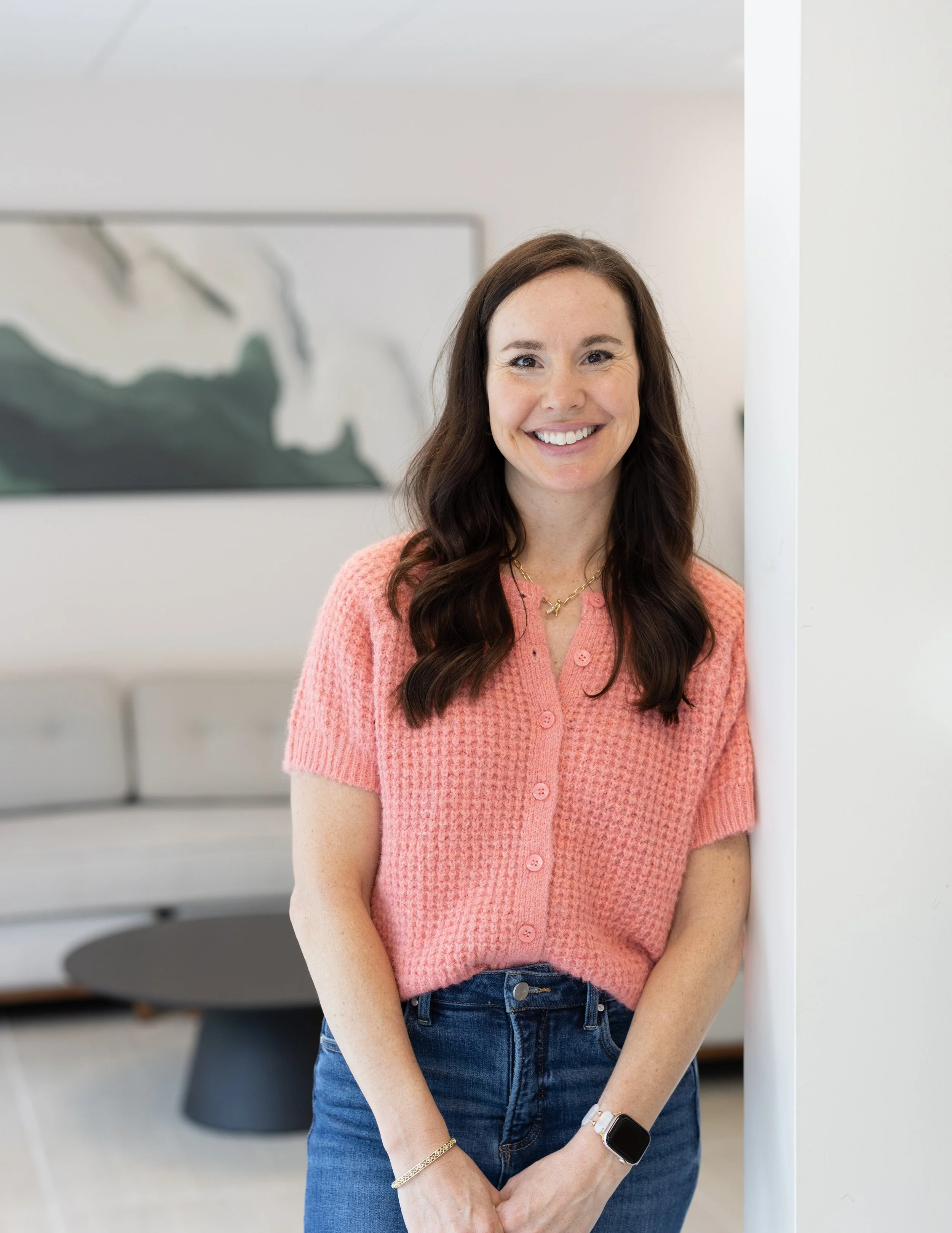 A woman with long brown hair smiling, standing indoors next to a white wall, wearing a pink cardigan, blue jeans, a gold necklace, a smartwatch, and a bracelet.