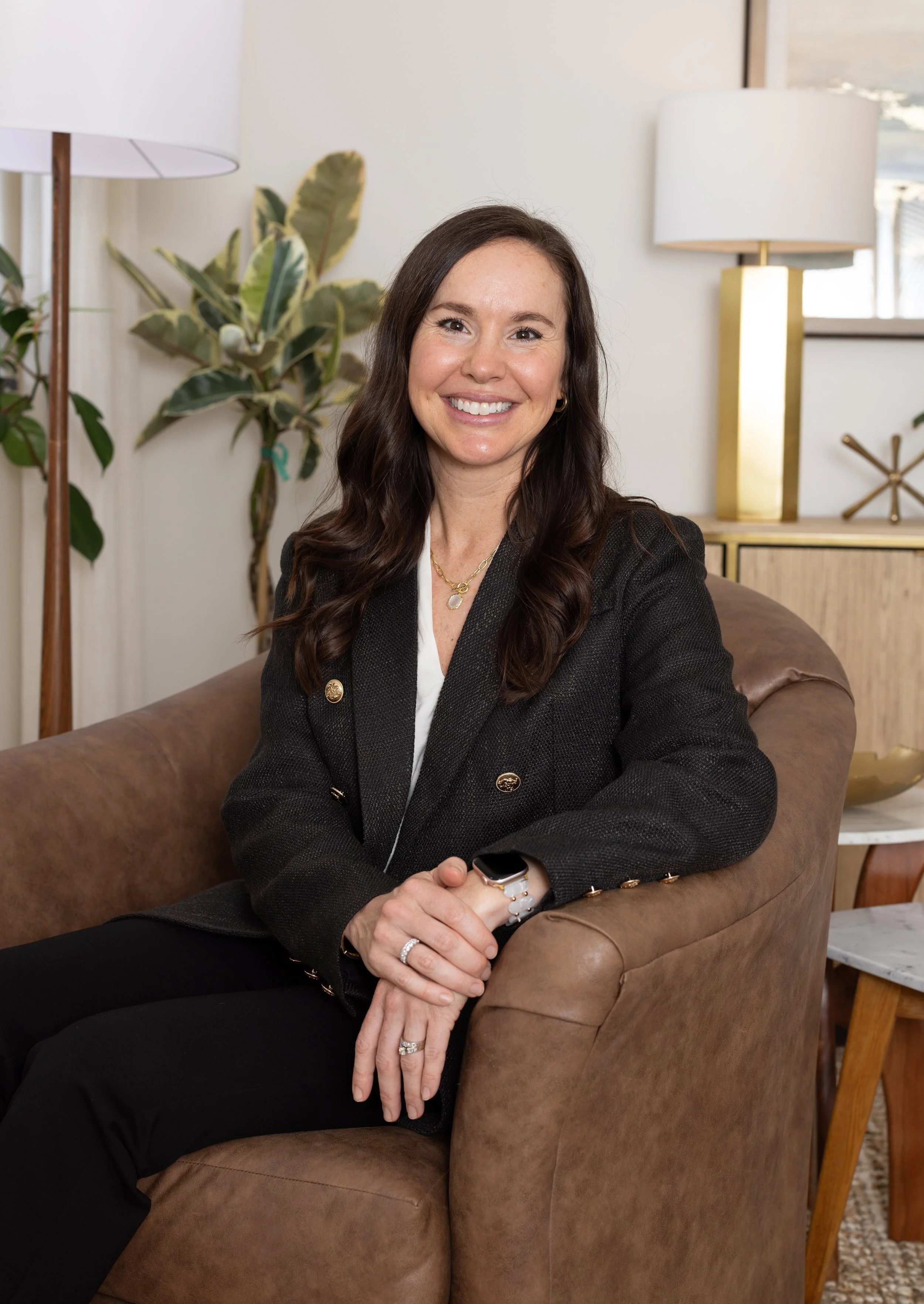 A woman with long dark hair sitting on a brown leather couch, smiling at the camera, wearing a black blazer, jewelry, and a smartwatch, in a living room with a plant and lamps in the background.