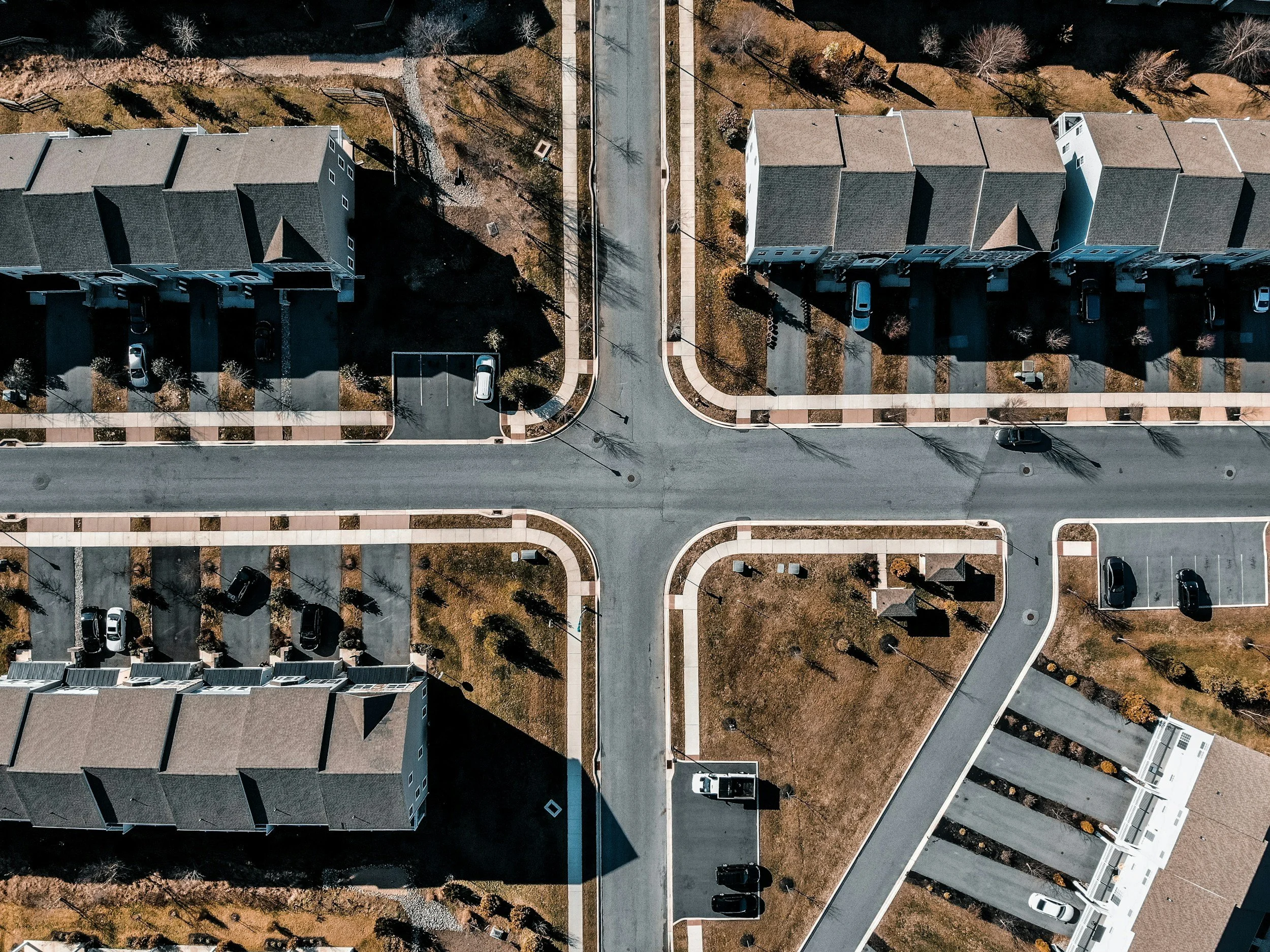 An aerial view of residential buildings with multiple cars parked in the driveways, surrounded by sidewalks, trees, and yards.