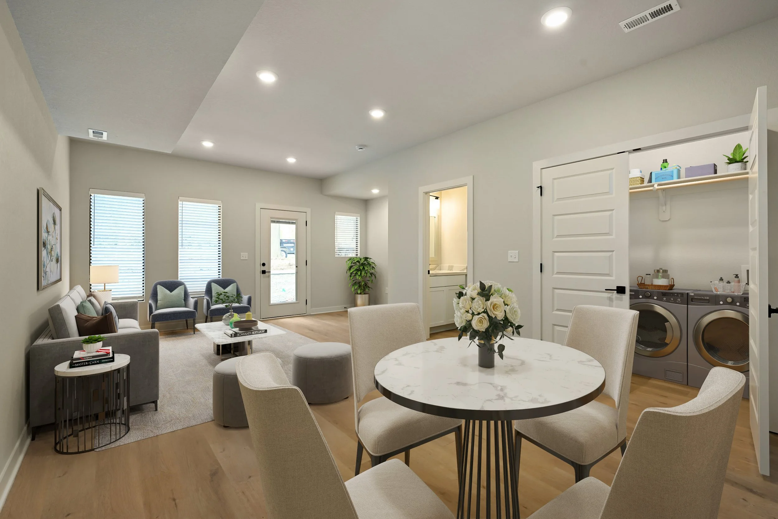 Living room with beige walls, three large windows with blinds, a white door, and a kitchen visible through an open doorway. The room features a gray sofa with pillows, a round dining table with six cream-colored chairs, a floral centerpiece, and a small side table. There is a washing machine and storage shelves in a nook.