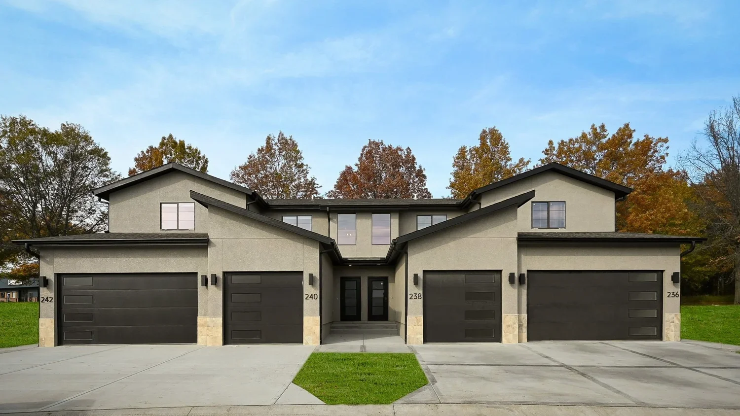 Modern townhouse with gray stucco exterior, black garage doors, and black front doors, flanked by two-car garages and surrounded by trees with autumn foliage.