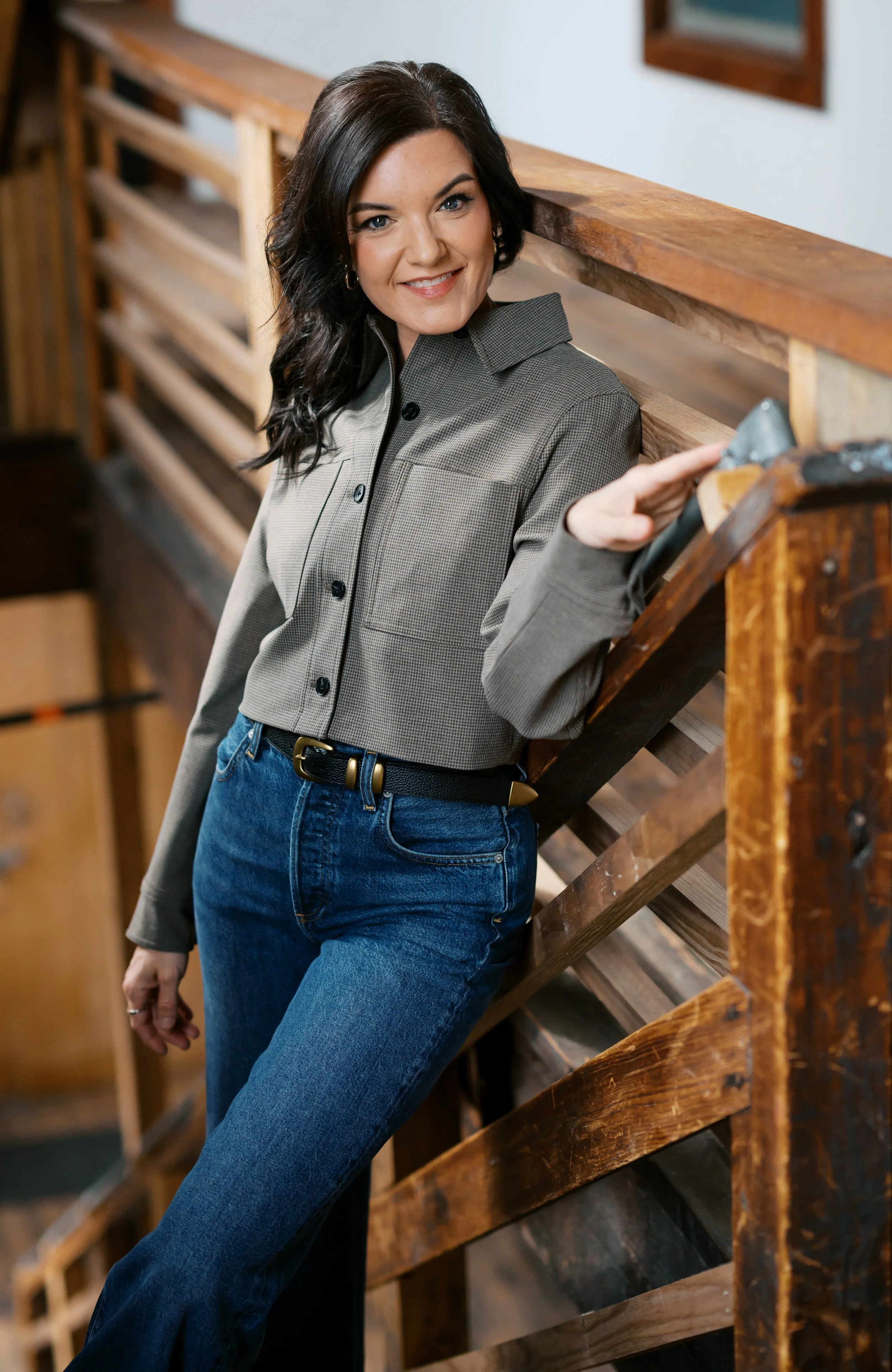 A woman with dark wavy hair, smiling, standing next to a wooden staircase, wearing a grey button-up shirt and blue jeans.