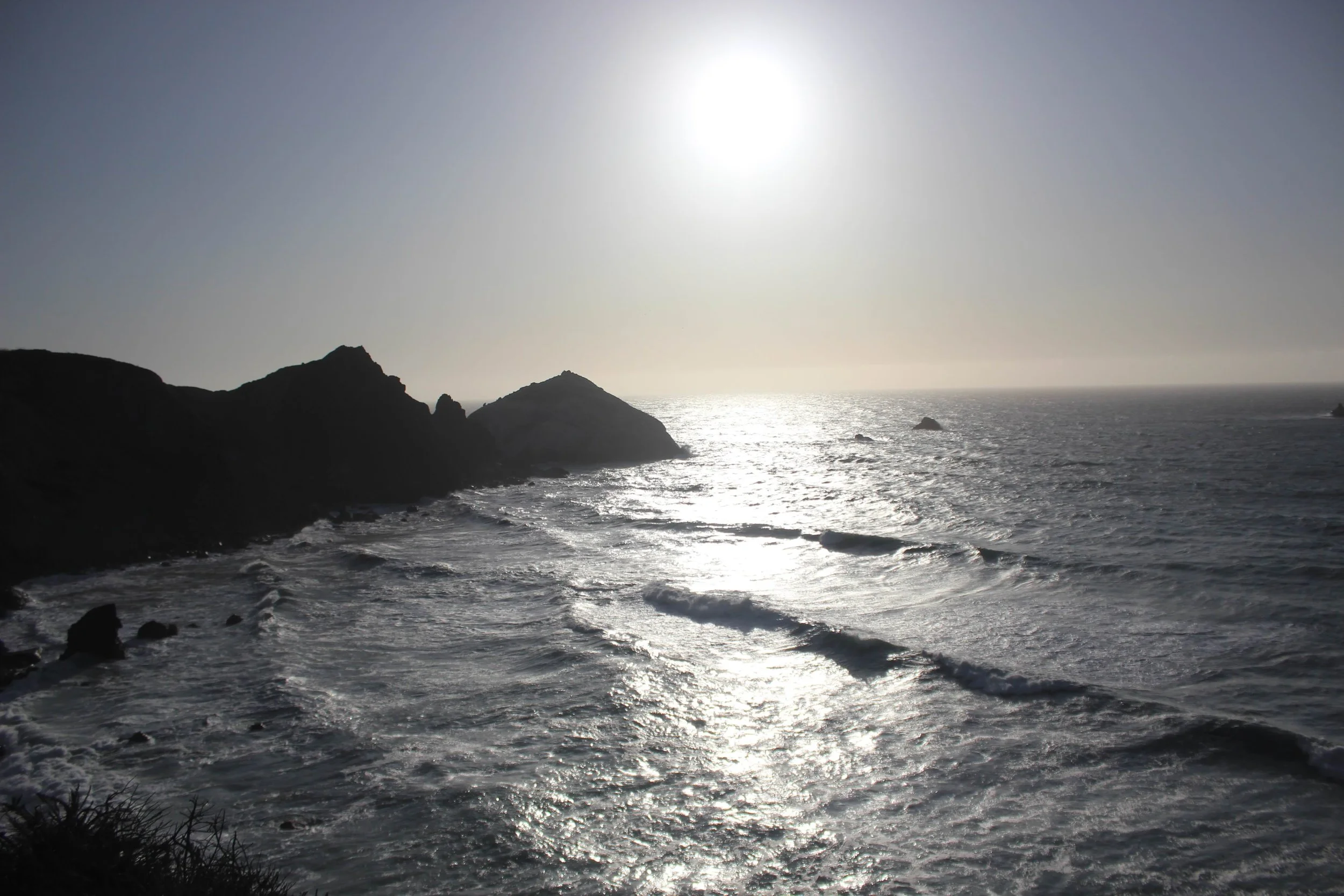Sunset over a rocky coastline with waves crashing against the shore, silhouetted cliffs in the foreground.