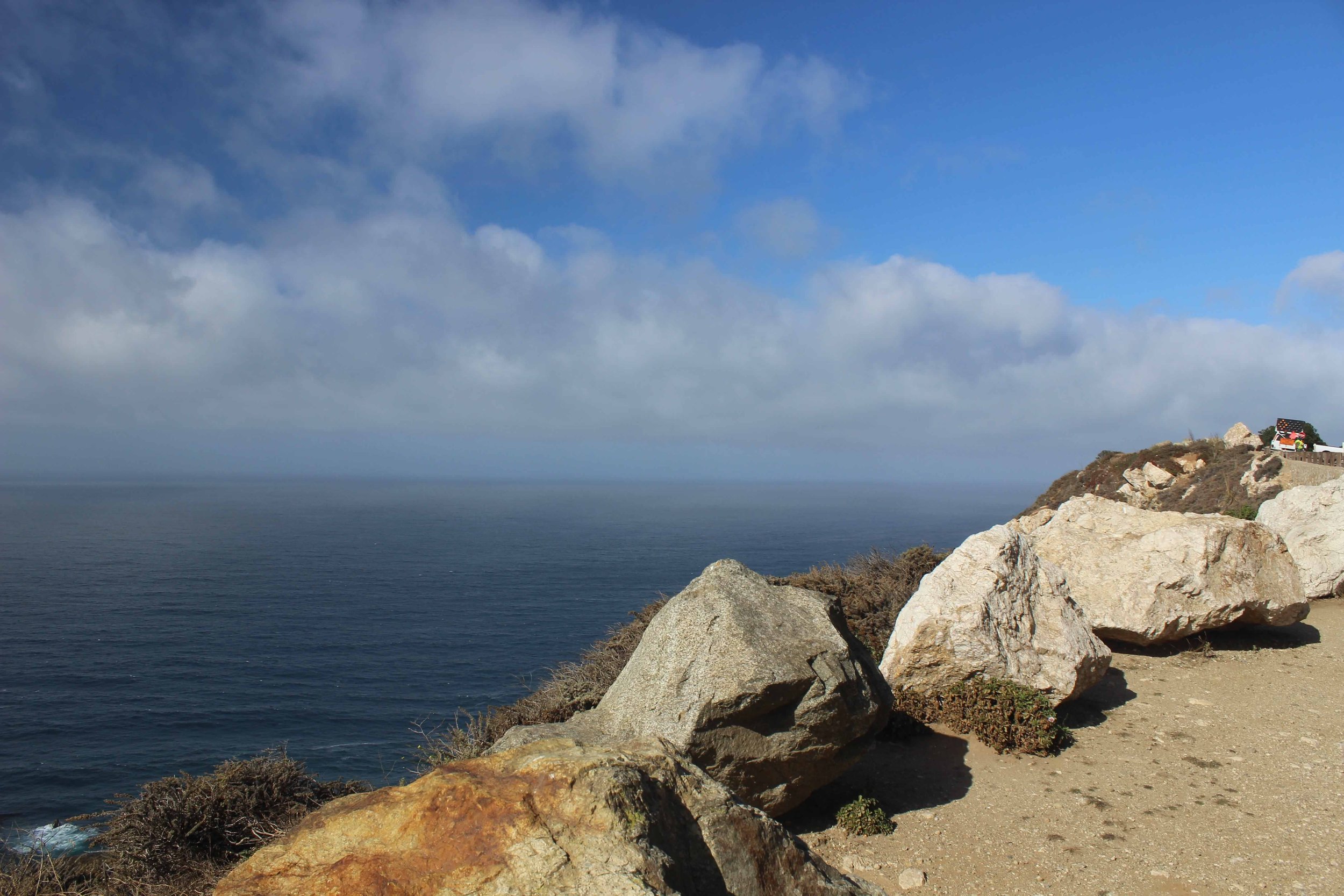 A rocky coastline with large rocks and a small house with an American flag on a cliff overlooking the ocean under a partly cloudy sky.