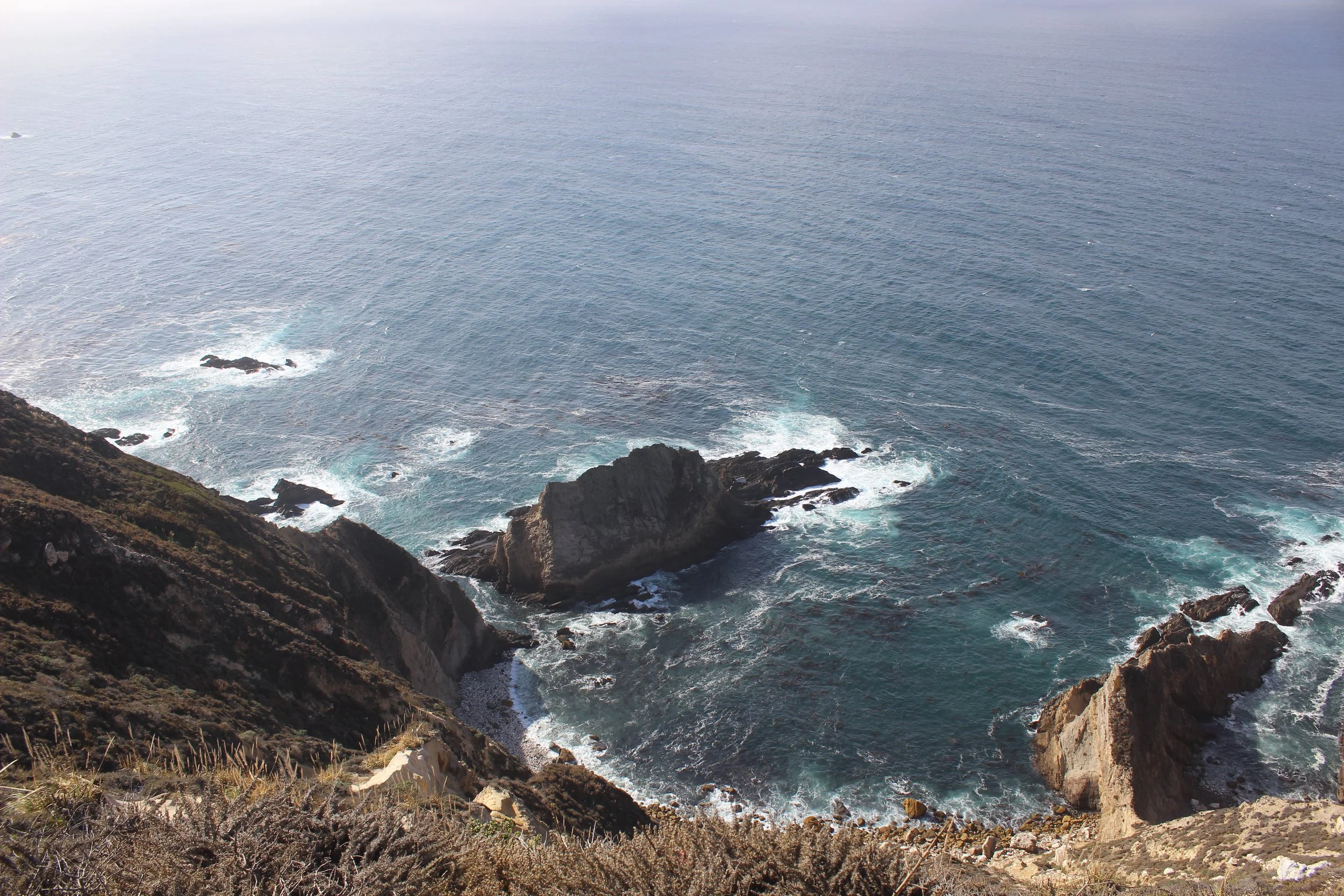 A rugged coastal scene with rocky cliffs and ocean waves crashing against the shoreline, viewed from a high vantage point.