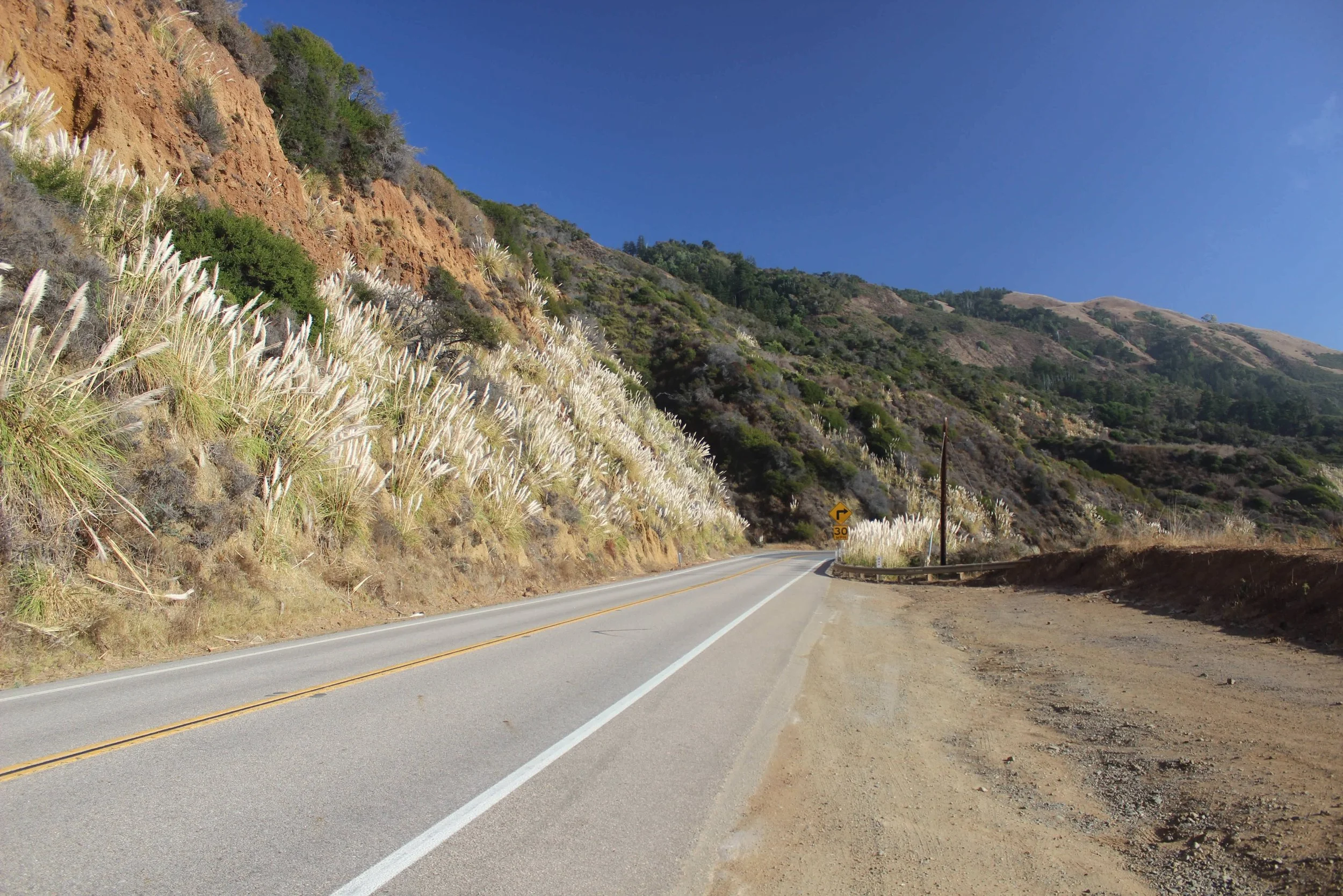 A winding mountain road with a 30 mph sign, flanked by steep hillside covered in grass and shrubs, under a clear blue sky.