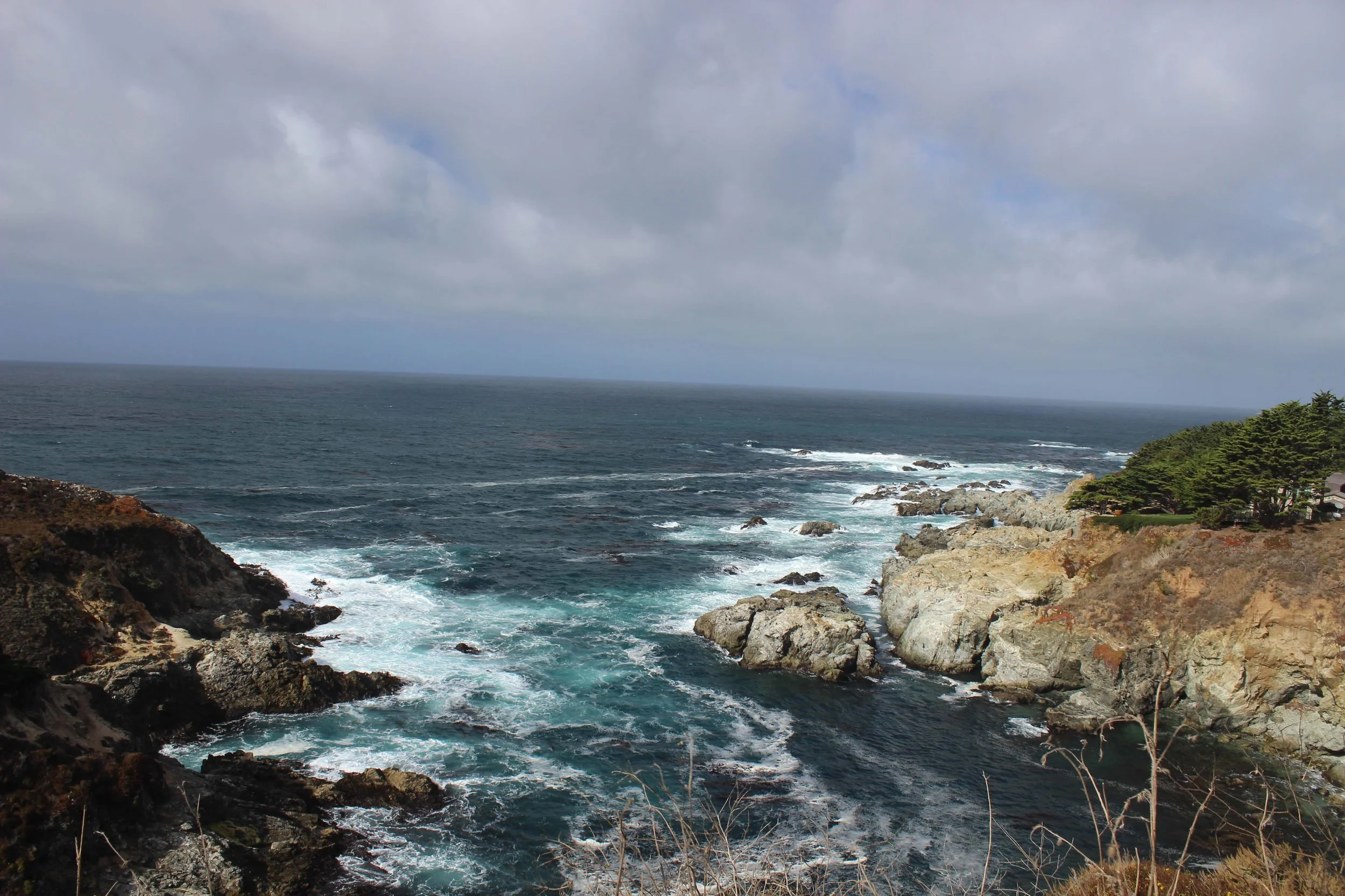 A rugged coastline with rocky cliffs and green shrubs, overlooking a choppy ocean with gray clouds overhead.