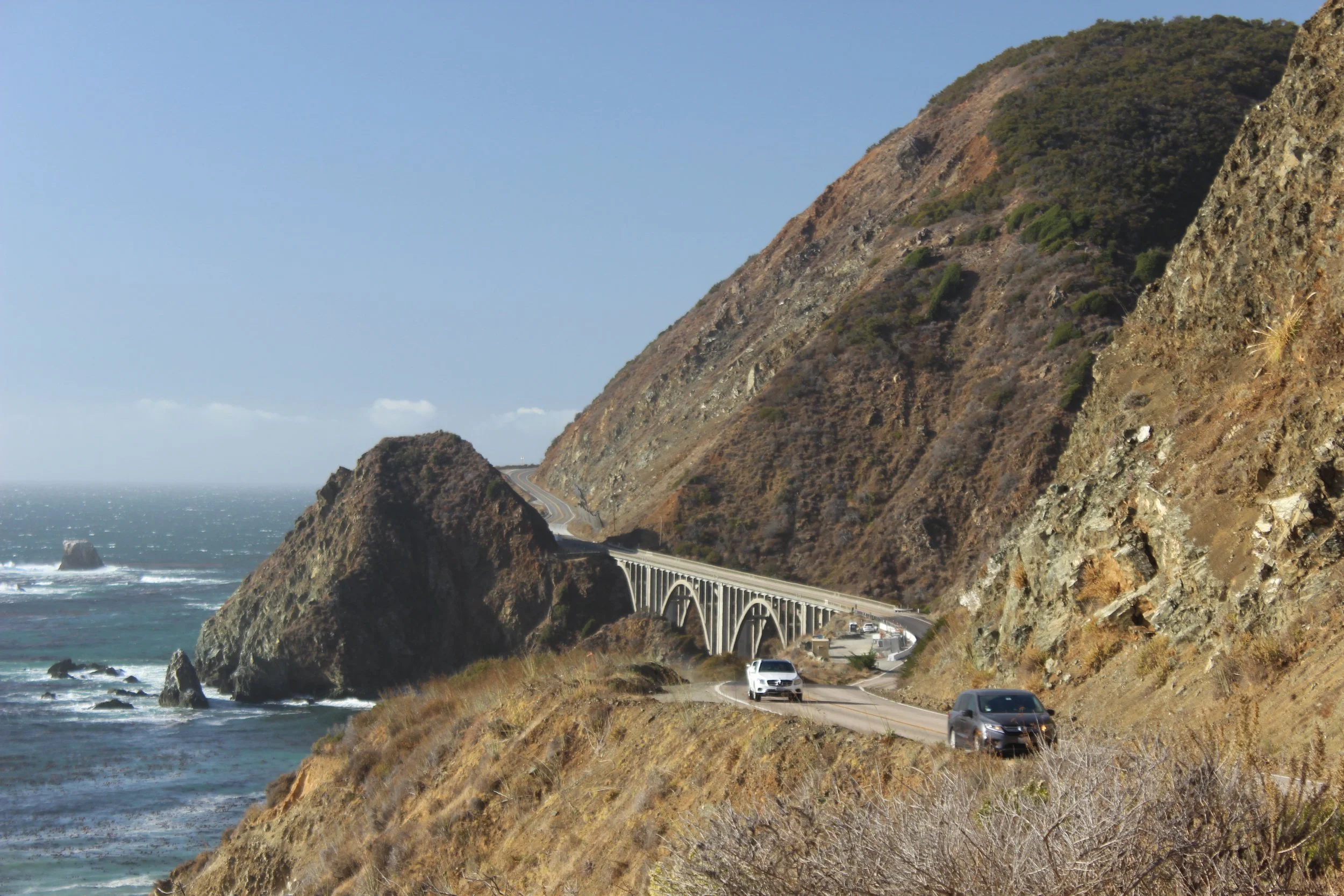 Coastal highway running along steep cliffs with cars driving by, ocean visible on the left, and a bridge supporting the highway.