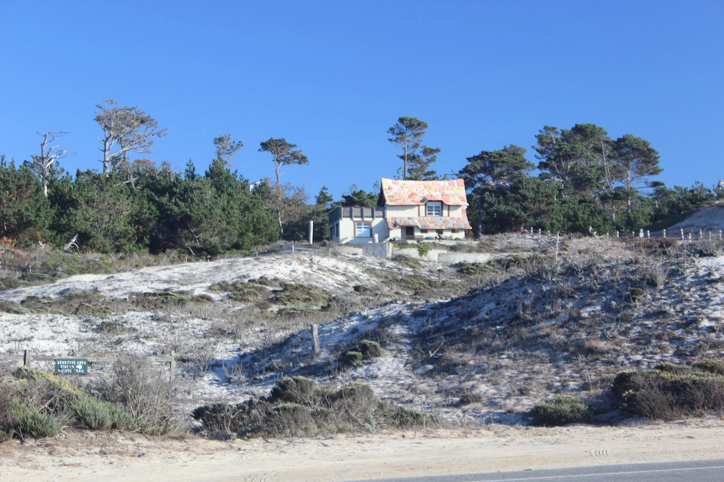 A house with a colorful shingle roof surrounded by trees on a hillside, with a dirt area and a sign that reads 'Sensitive Area Stay on Trail' in the foreground.