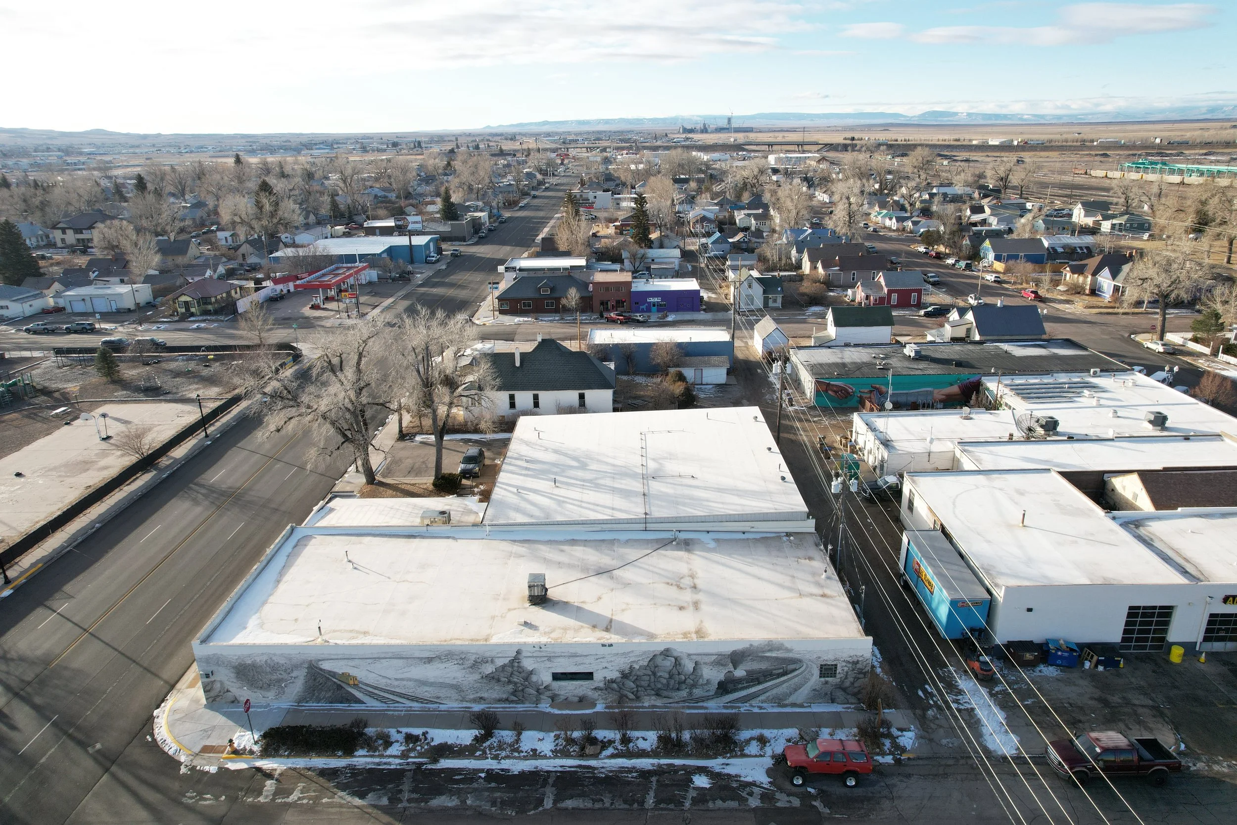 Aerial view of a small town with residential houses, commercial buildings, and streets, with a mountain range in the distance and some snow on the rooftops and ground.