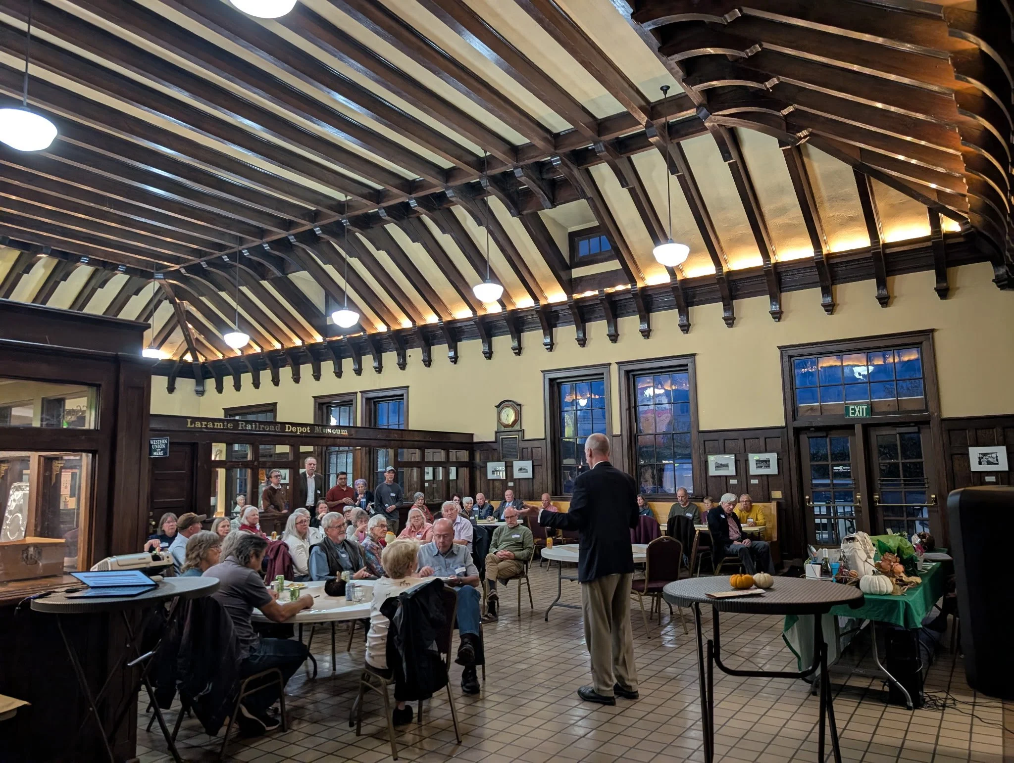 Indoor gathering at a historical site, possibly a museum or meeting hall, with a high, wooden beamed ceiling, large windows, and a group of people seated at tables and listening to speakers.