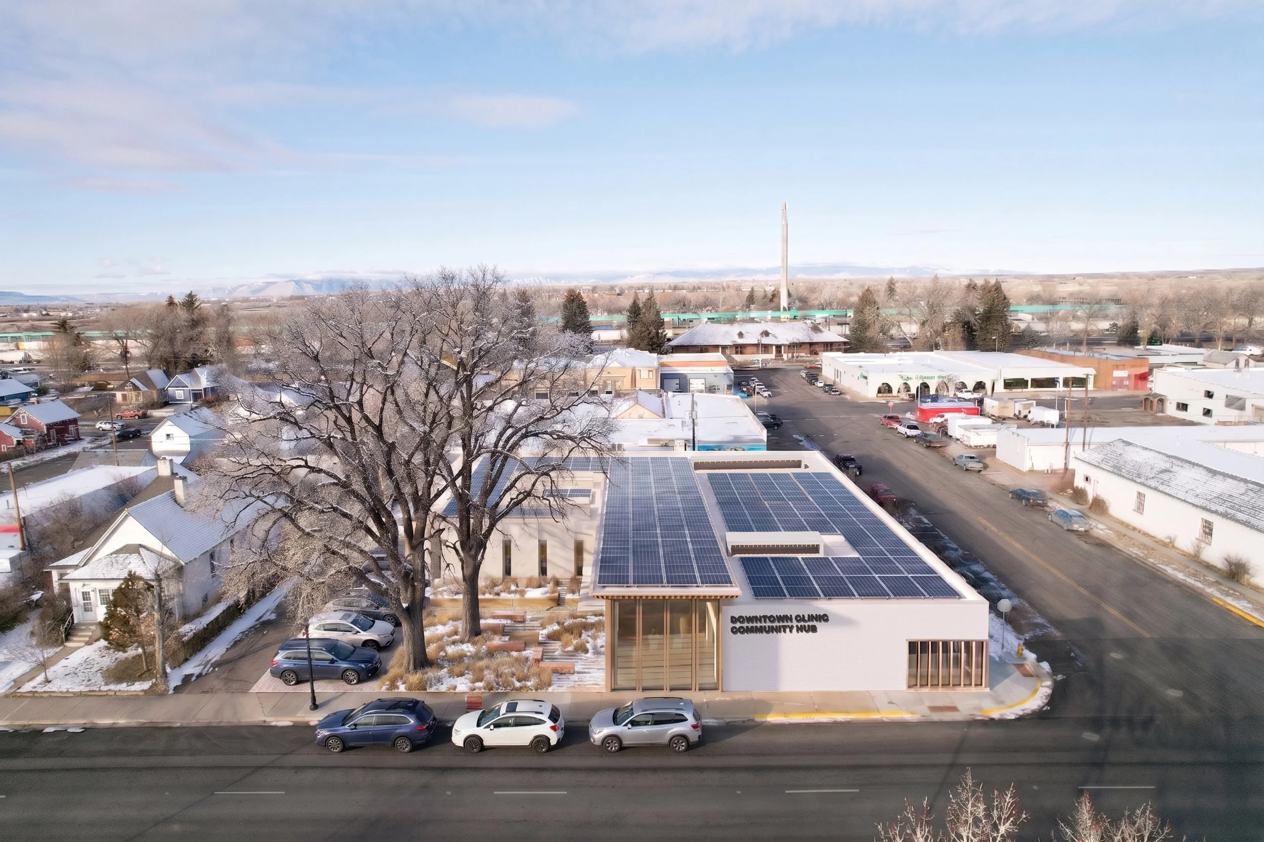 Aerial view of a snowy town with houses, trees, a building labeled 'Downtown Clinic Community Hub' with solar panels on the roof, and a parking lot in front.