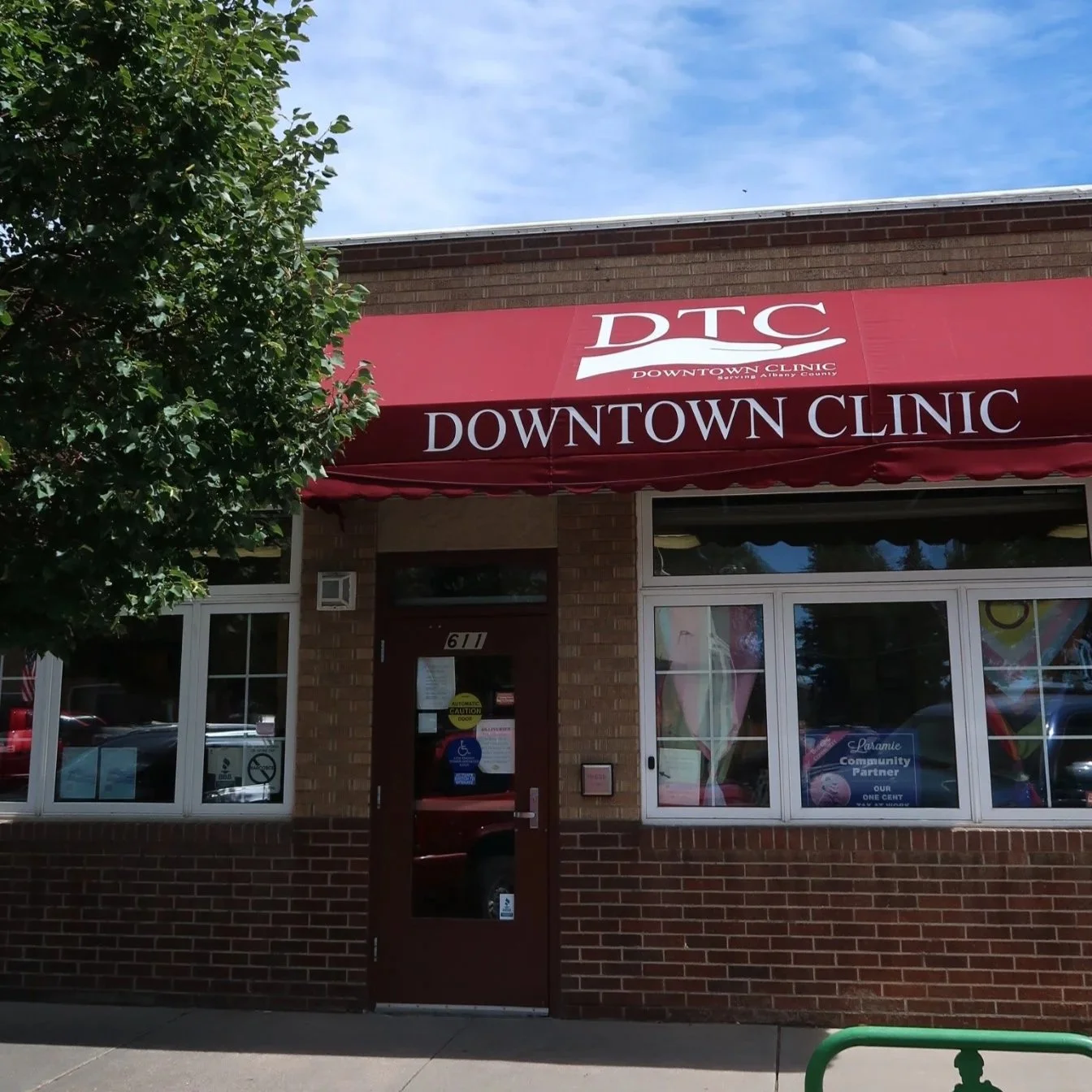 The exterior of a brick building with a red awning that reads 'DTC DOWNTOWN CLINIC'. There is a tree on the left and some parked cars reflected in the windows.