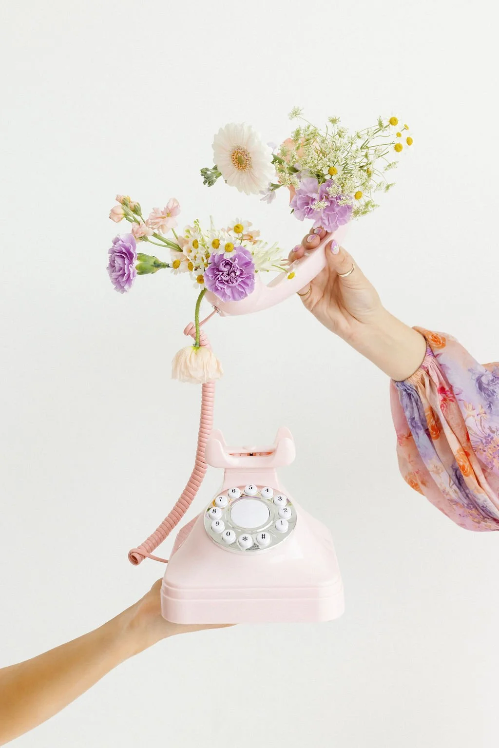 Two people hold a vintage pink rotary phone with a bouquet of colorful flowers emerging from the receiver against a plain white background.