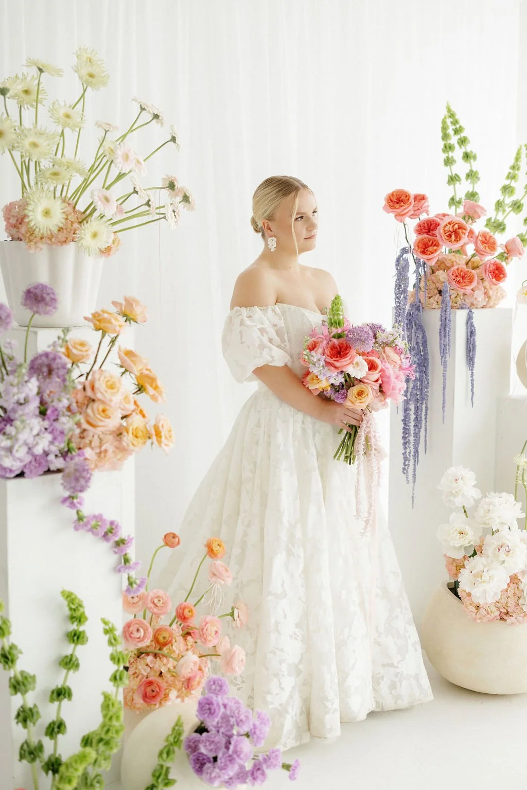 A woman in a white gown holding a colorful bouquet of flowers, surrounded by various pastel-colored floral arrangements in a bright, airy setting.