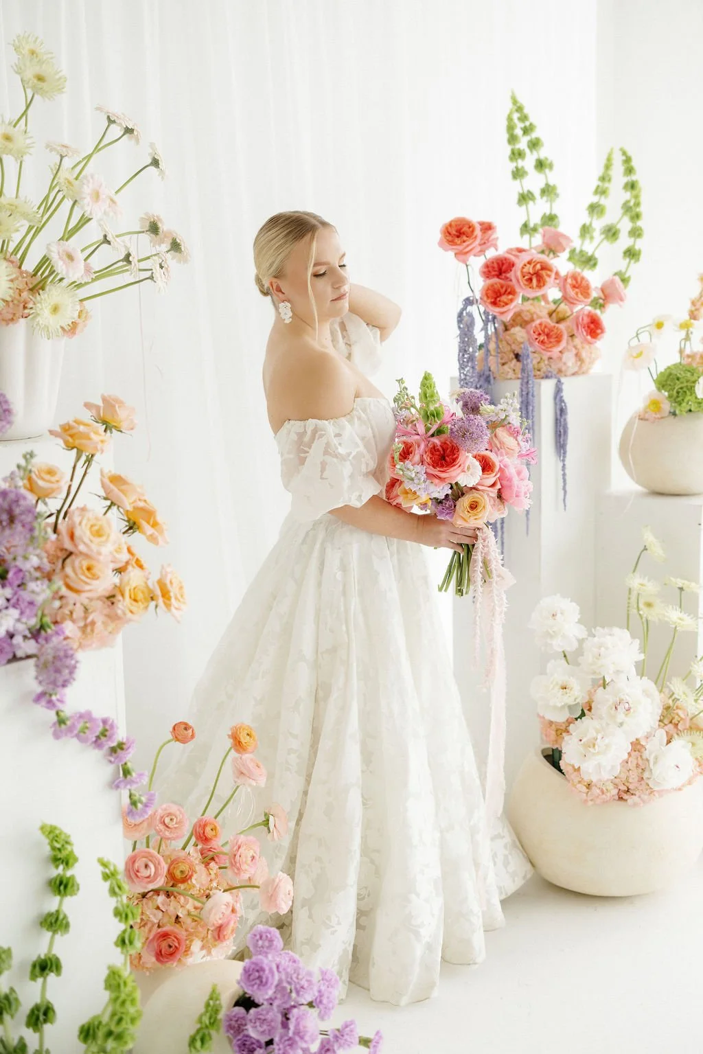 A woman in a white floral wedding dress holding a bouquet of pink and purple flowers, surrounded by various flower arrangements in white vases and pots in a bright, airy setting.