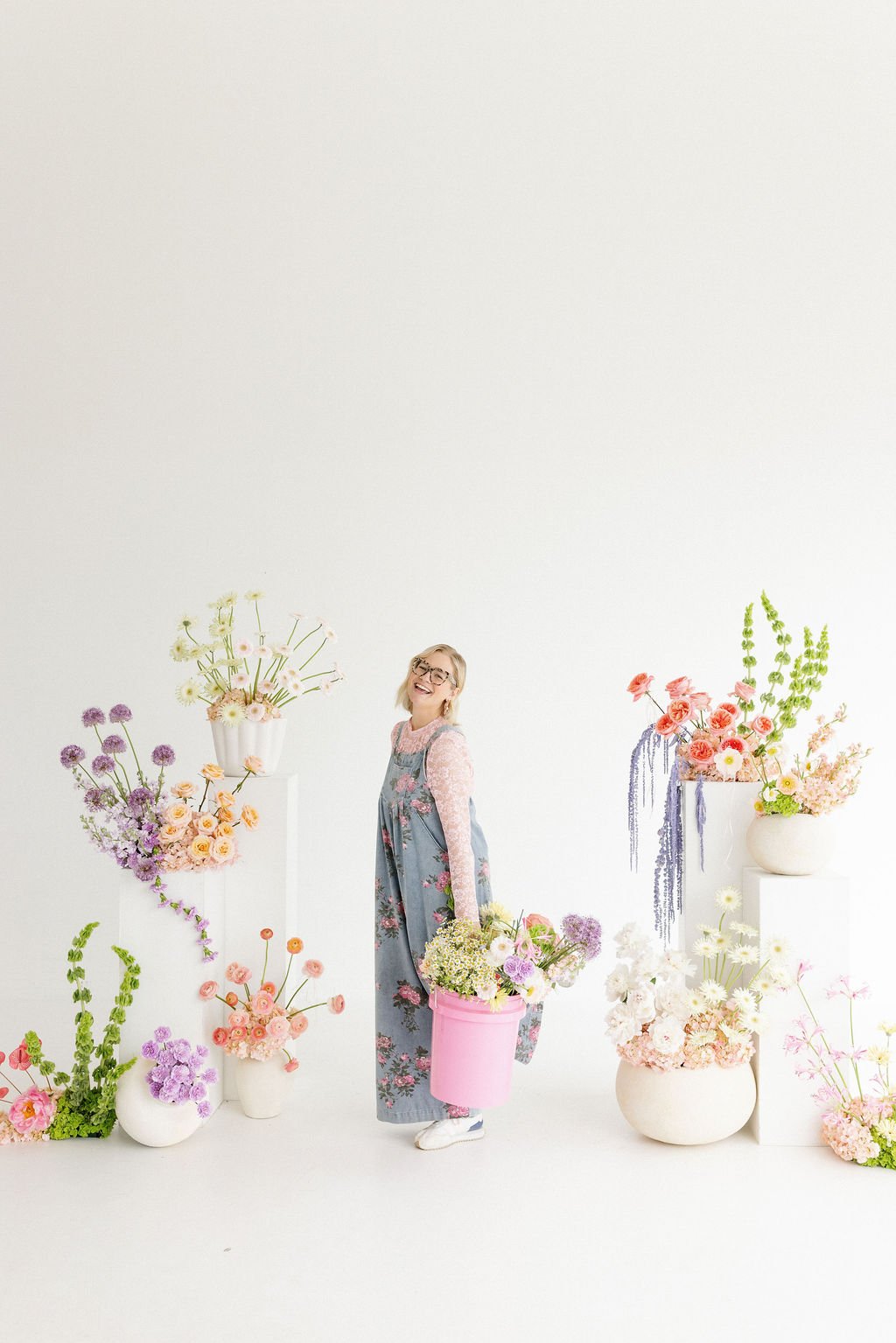 Woman smiling and holding a pink bucket filled with flowers, surrounded by colorful flower arrangements in vases on white pedestals and the floor, set against a plain white background.