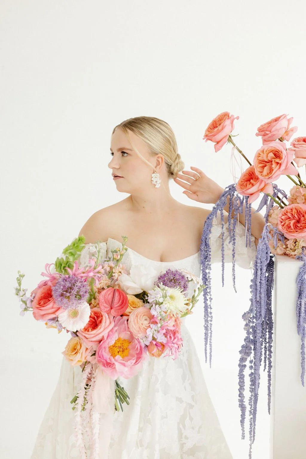 A bride in a white dress holding a bouquet of pink, purple, and white flowers, with more flowers and hanging purple blossoms on a white pedestal.