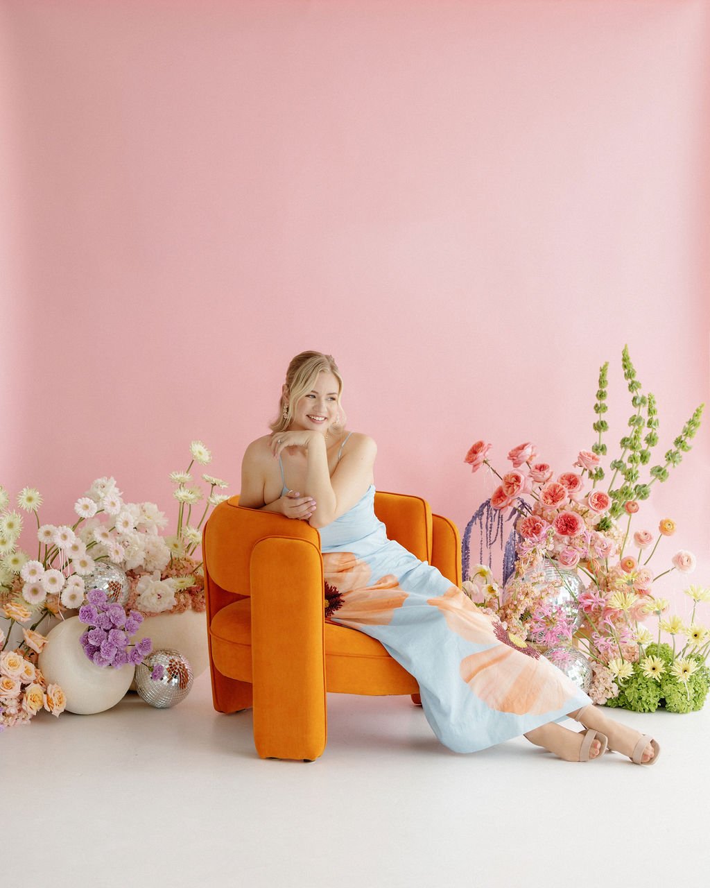 A woman sitting on an orange chair amidst pink and purple flowers against a pink background, smiling and resting her chin on her hand.
