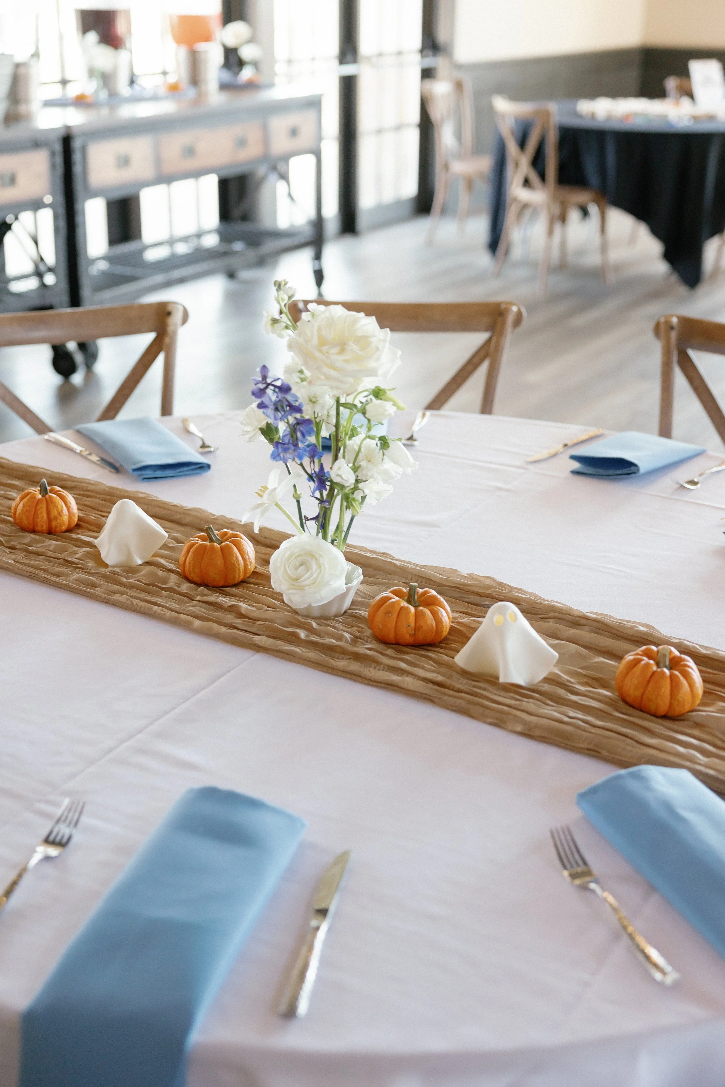 A decorated dining table with a white tablecloth, blue napkins, and silverware. The table has a tan table runner, small pumpkins, and a floral centerpiece with white and purple flowers. In the background, there are other tables and chairs in a bright, airy room.
