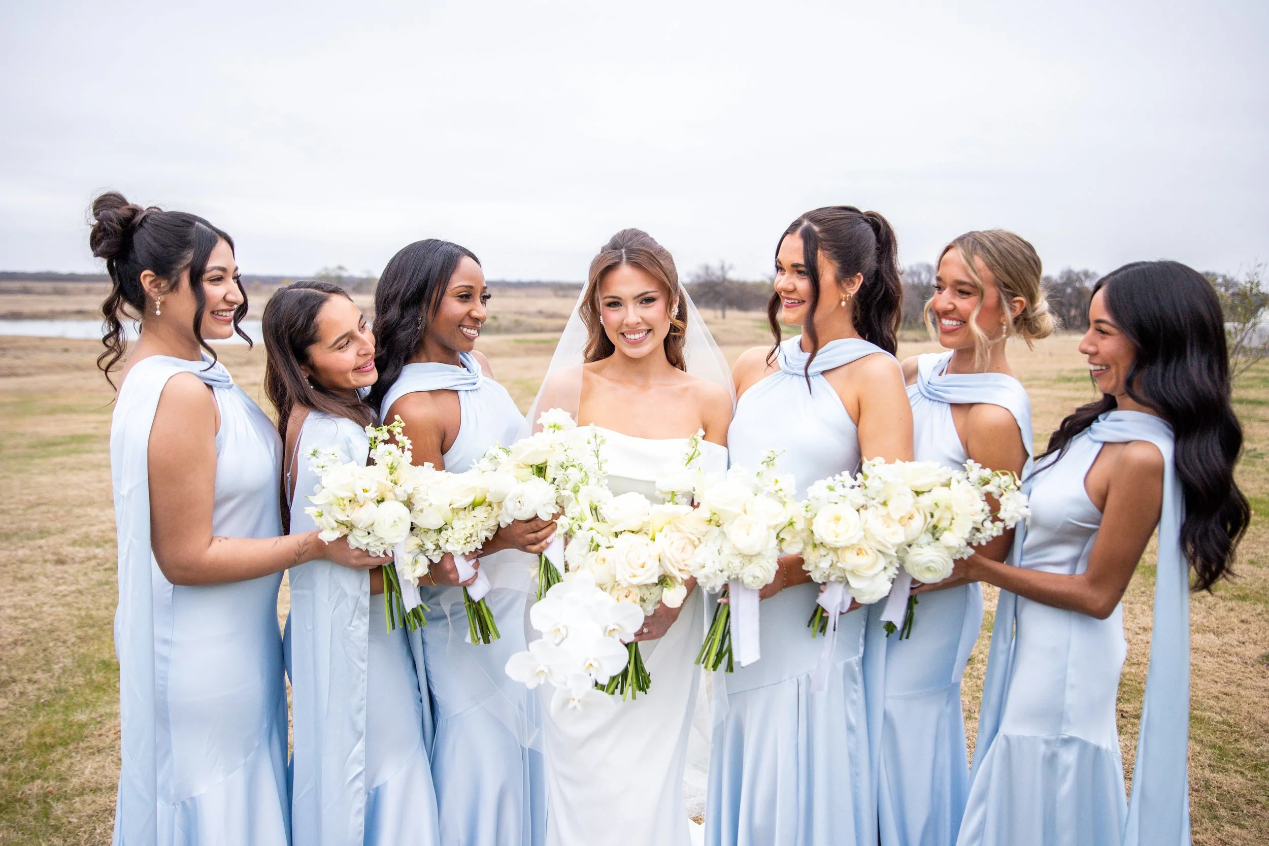 A bride and six bridesmaids standing outdoors on a cloudy day, all dressed in matching light blue dresses, smiling, and holding bouquets of white flowers.