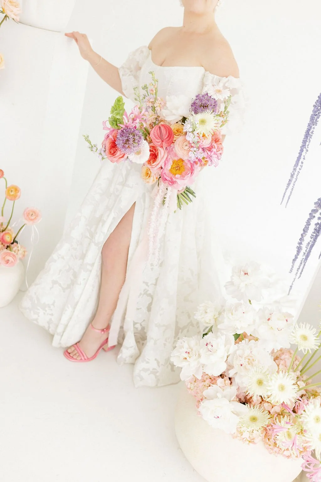 Woman in a white off-shoulder lace dress with a slit, holding a large bouquet of colorful flowers, in a bright, minimalistic room with white walls and floor, decorated with pink and white roses and other flowers.