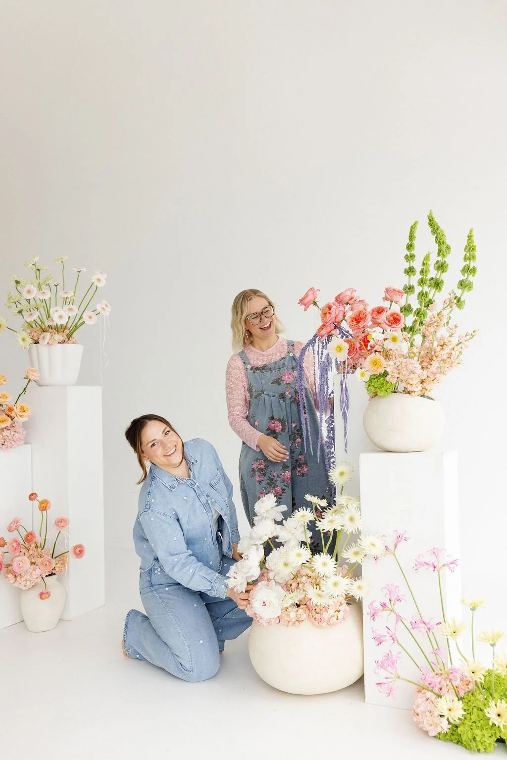 Two women in denim outfits smiling among large white vases filled with colorful flowers, in a minimalistic white studio setting.