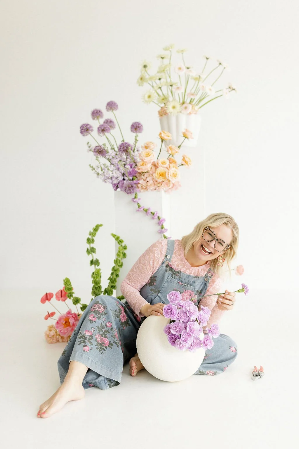 A woman with blonde hair and glasses smiling, sitting barefoot on the floor holding a purple flower. She is surrounded by various colorful flowers and a white vase. The background is white with a tall arrangement of flowers hanging on the wall.