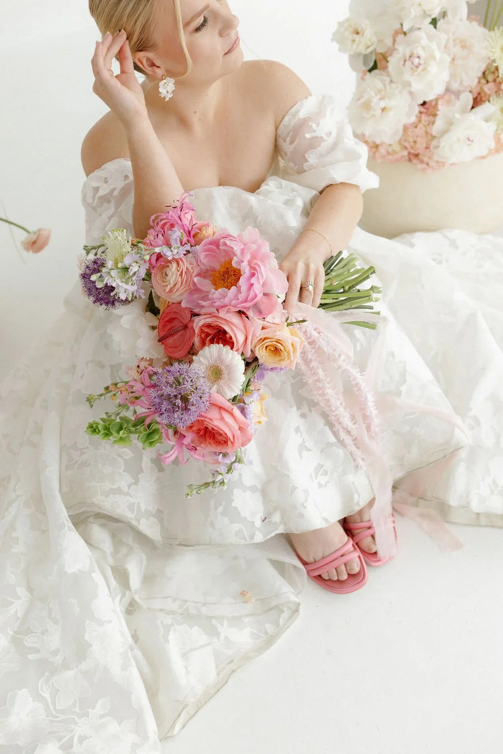 A woman in a white floral wedding dress holding a large bouquet of pink and peach flowers, sitting on the floor with her eyes closed and head tilted to the side, wearing pink sandals and floral earrings.