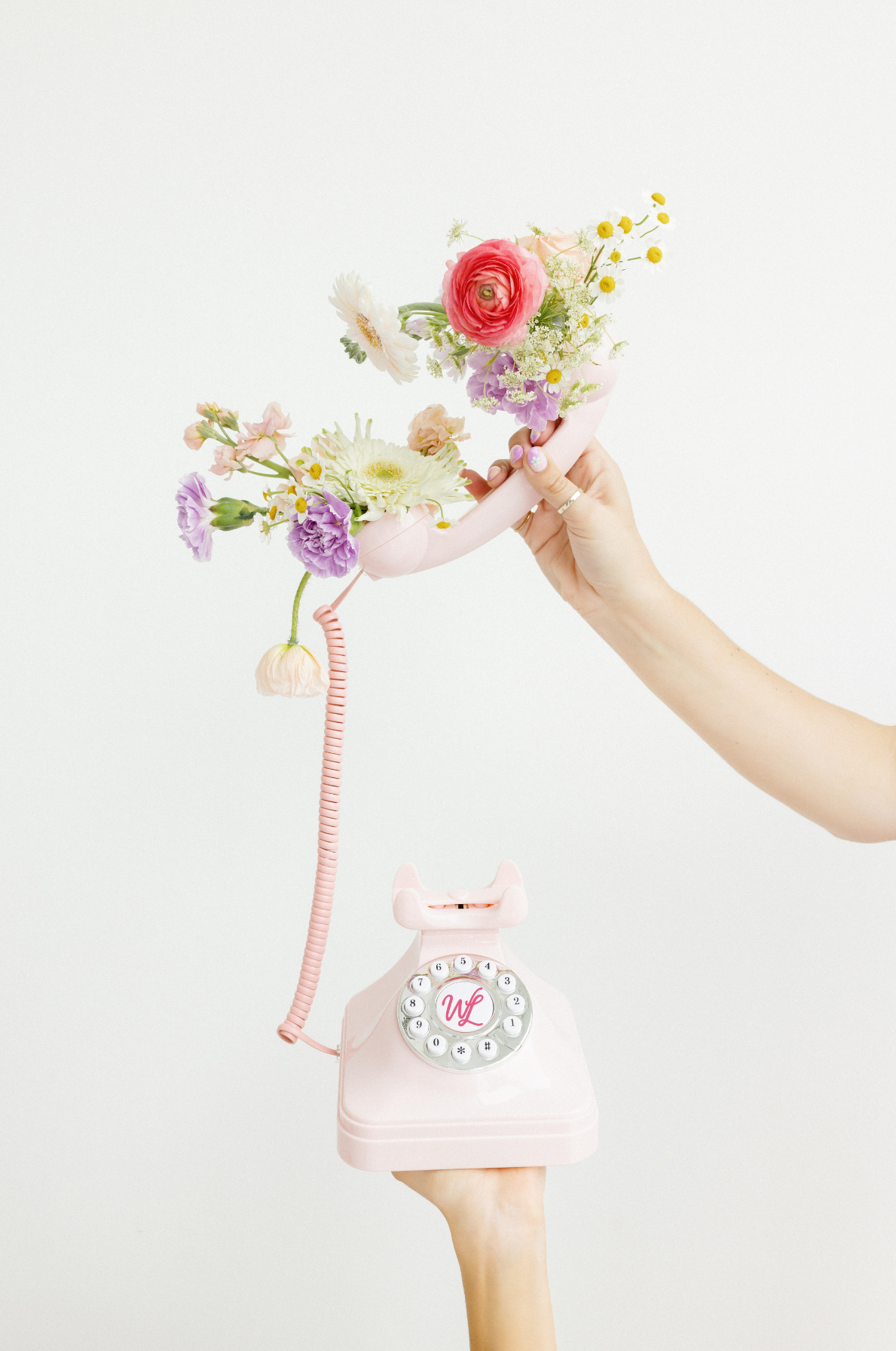 A hand holding a pink vintage rotary telephone with the receiver attached to a bouquet of colorful flowers, including pink, white, and purple blooms, and a hand holding the other side of the phone is visible.