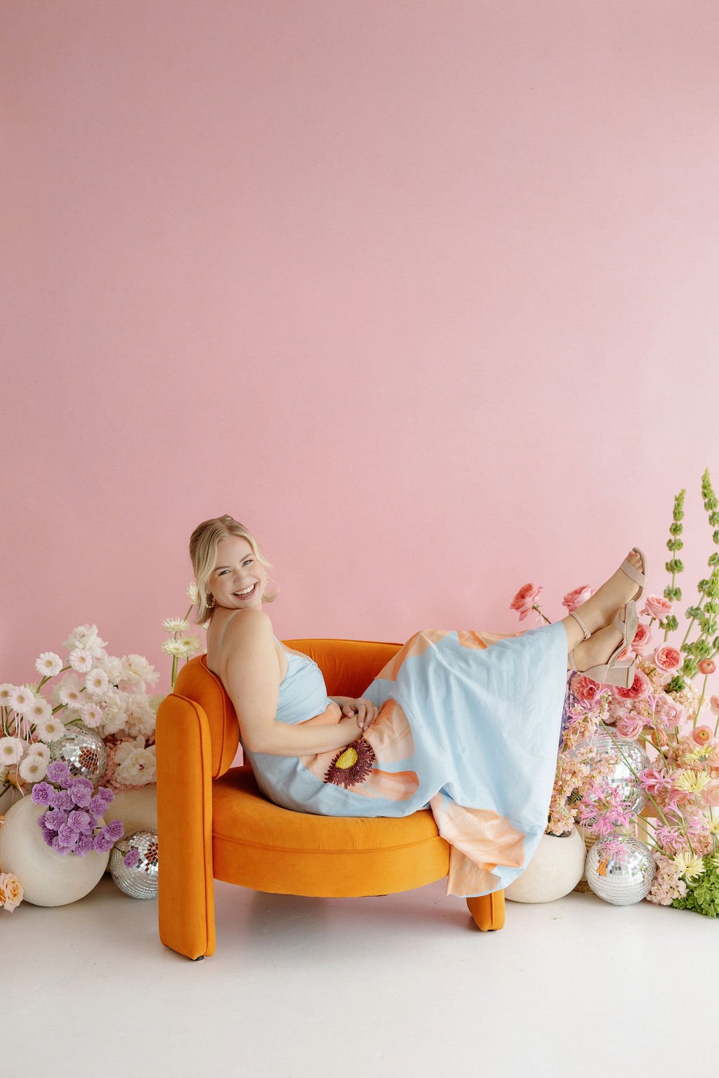 A woman with blonde hair, smiling and laughing, reclining on an orange velvet sofa surrounded by pink, purple, and white flowers, with a pink background.