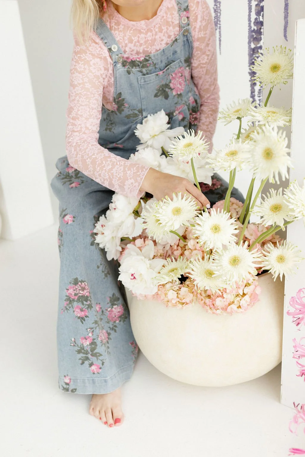 Person arranging white and pink flowers in a large white pot, dressed in pink lace top and floral overalls.