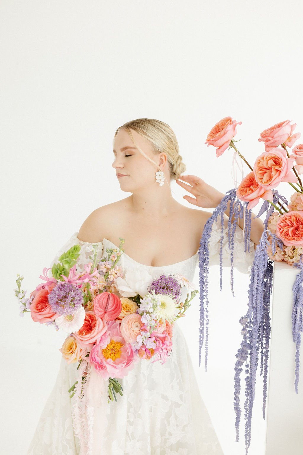 A woman in a white off-shoulder dress holding a bouquet of pink, purple, and white flowers, with a woman adjusting her earring, surrounded by colorful floral arrangements and purple hanging flowers, against a white background.