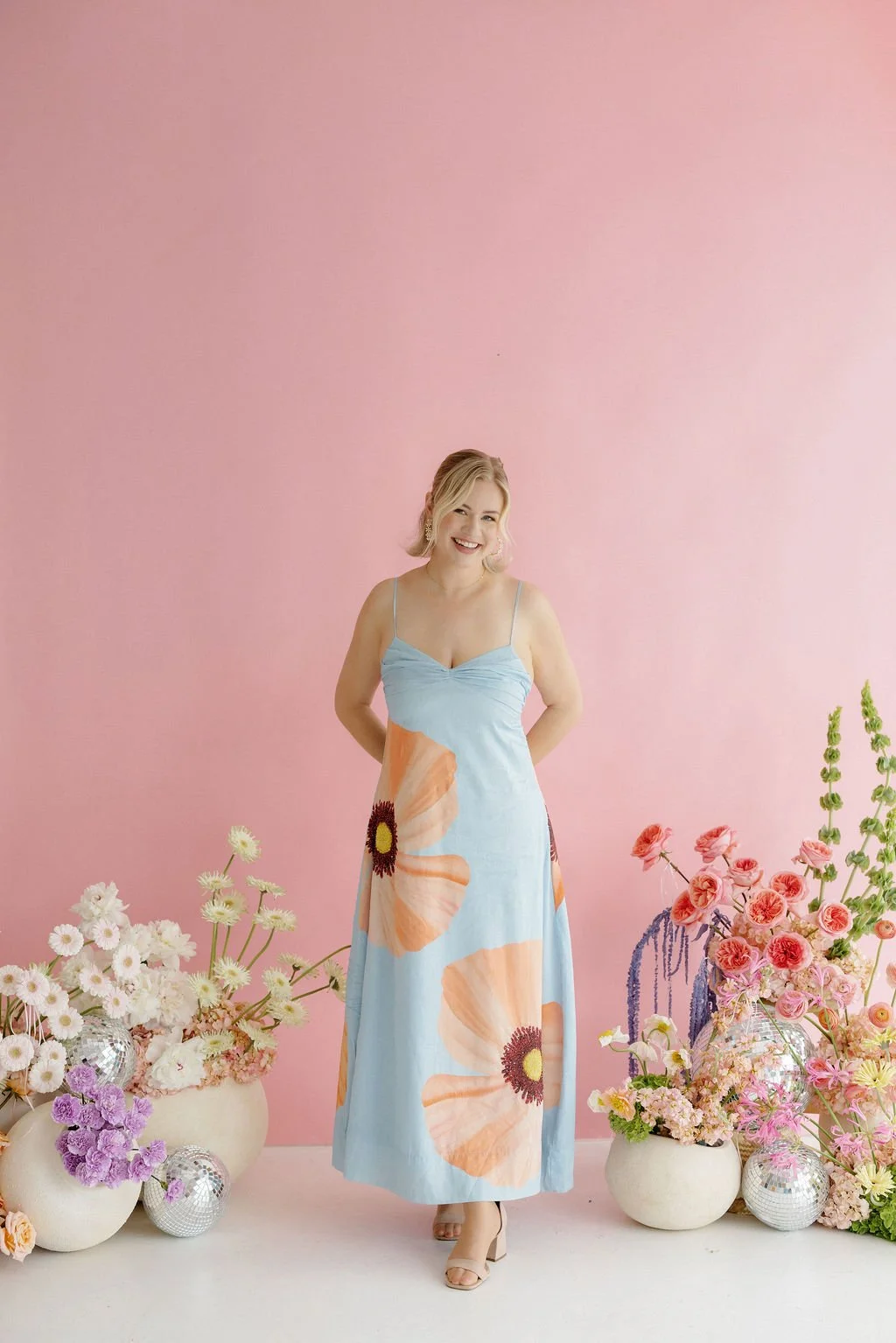Woman standing in front of a pink wall, wearing a floral dress and surrounded by vases of flowers and decorative disco balls.