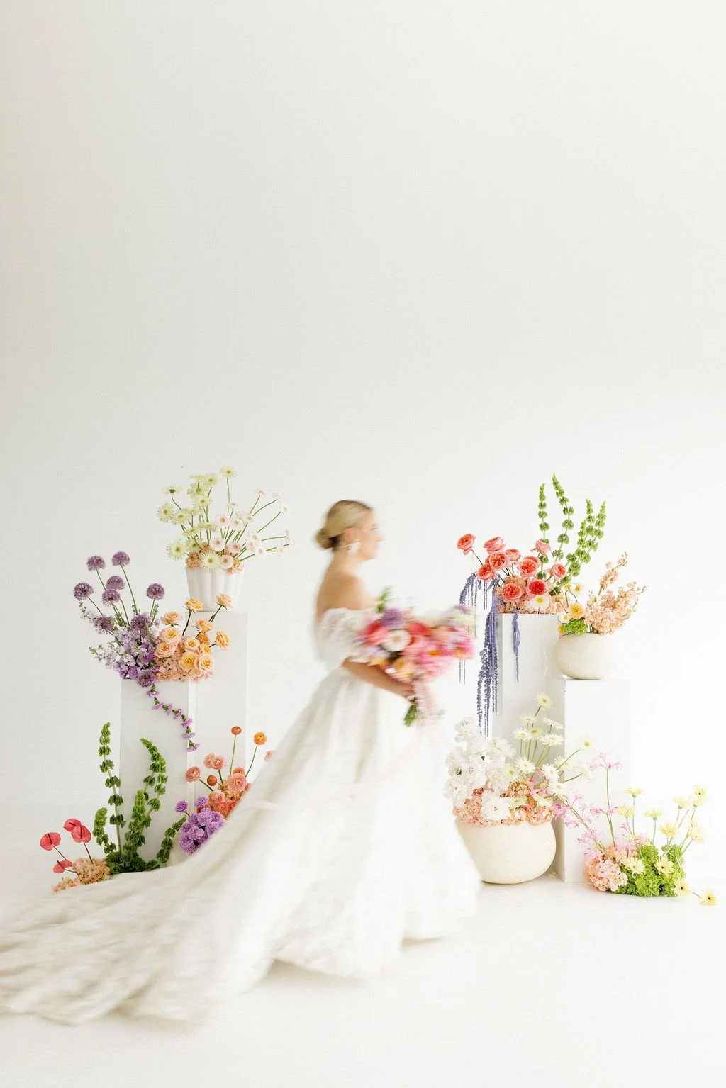 Bride in white wedding dress walking past colorful flower arrangements in white vases against a plain white background.
