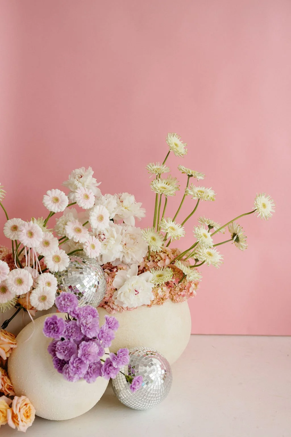 Pink background with white and purple flowers in vases, decorated with small disco ball ornaments.