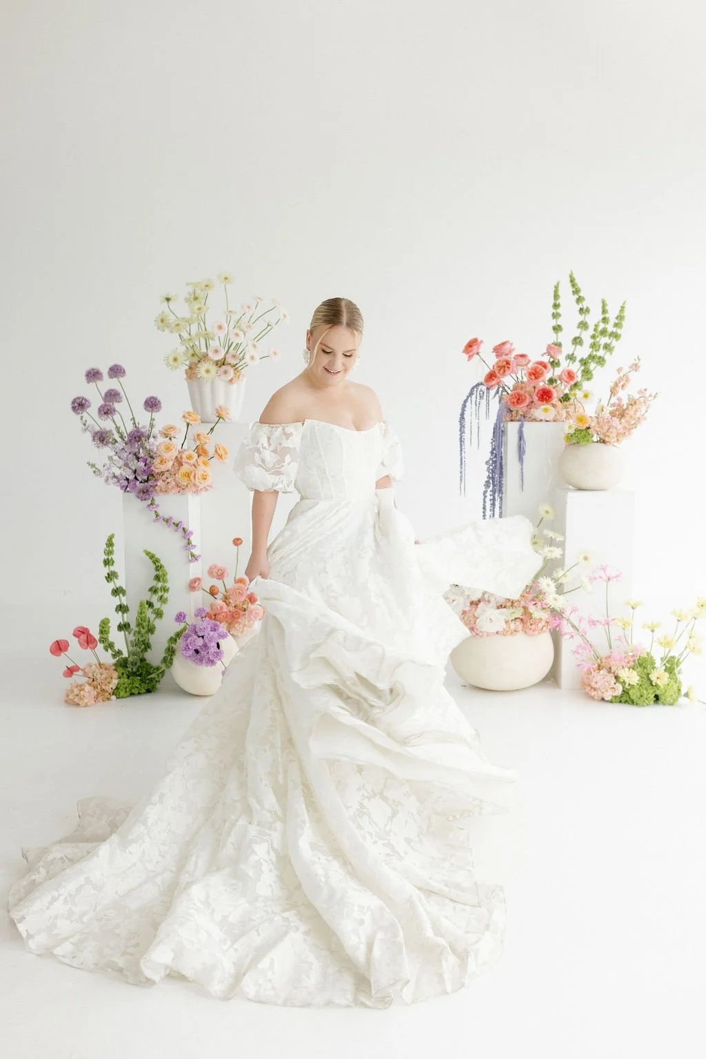 A woman in a white wedding dress surrounded by colorful flowers in vases on white pedestals, standing against a white background.