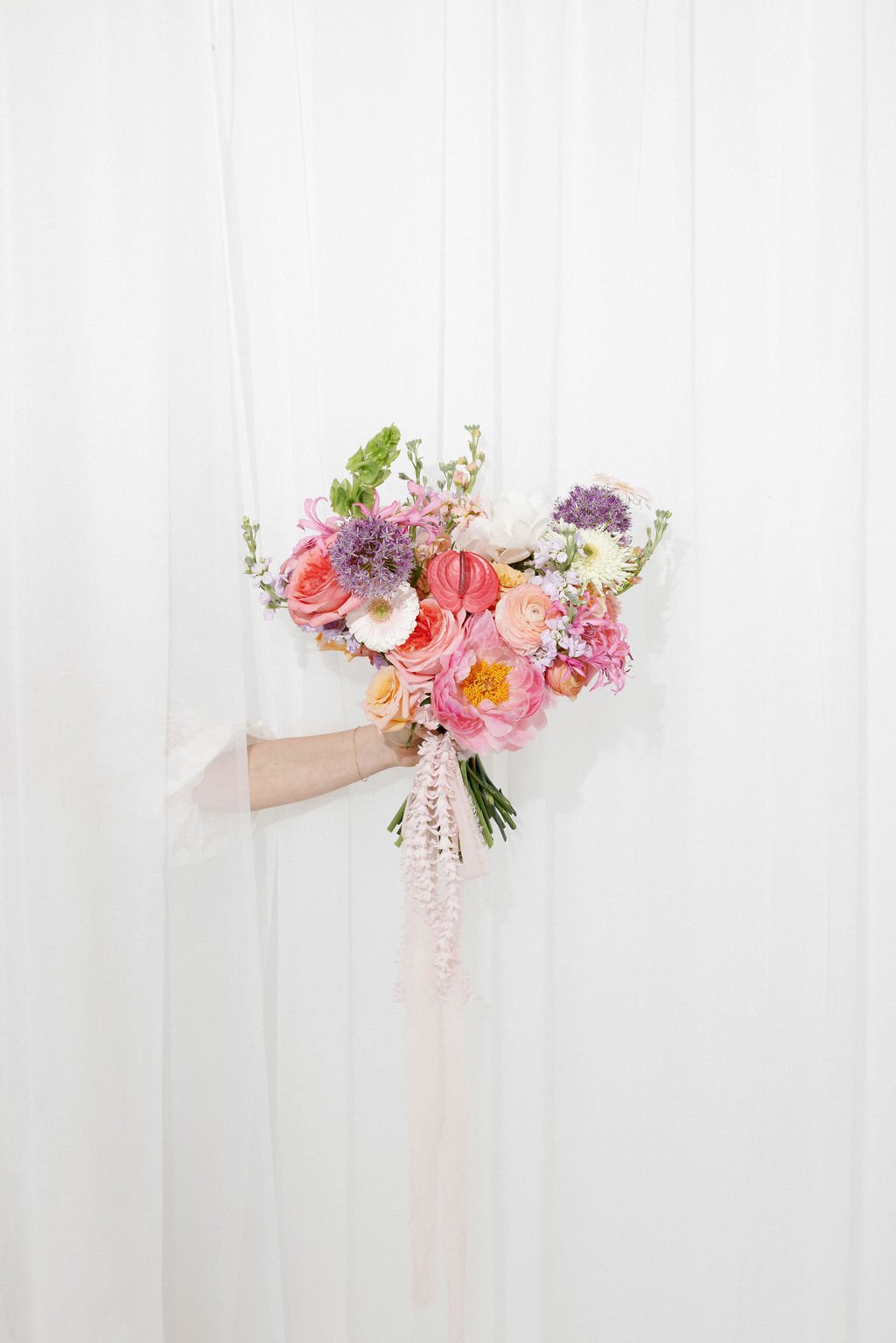 A bouquet of colorful flowers including pink roses, purple alliums, white daisies, and other blooms, being held in front of white paneled curtains.