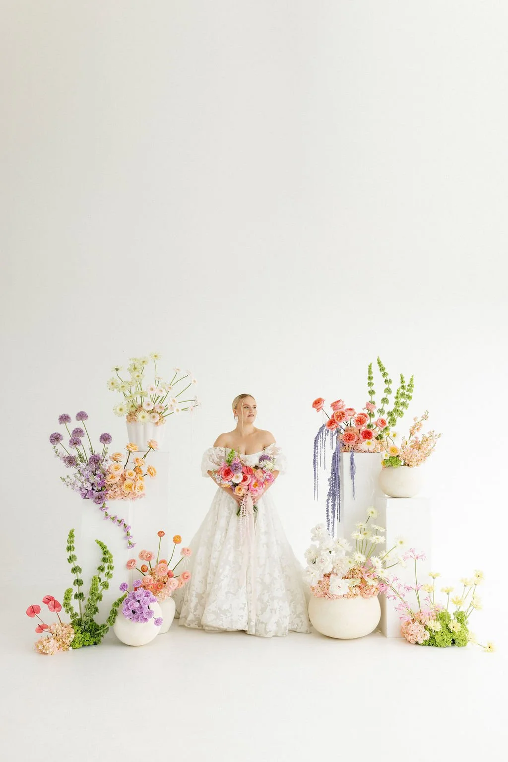 A woman in an off-shoulder wedding dress holding a large bouquet of colorful flowers, surrounded by tall flower arrangements in white vases against a plain white background.