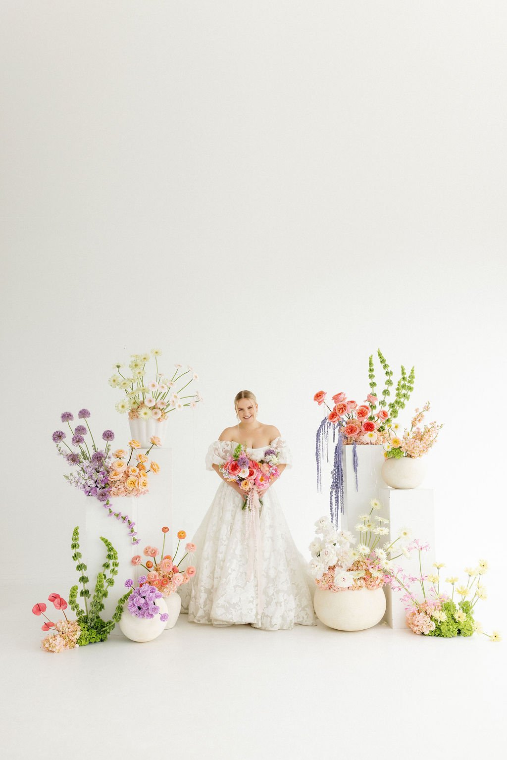 A woman in a wedding dress holding a bouquet of pink and purple flowers surrounded by large white vases filled with colorful flowers, set against a white background.