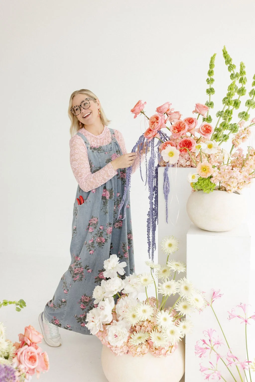 A woman with blonde hair, glasses, and a floral jumpsuit arranging pink, purple, and white flowers in a white vase, surrounded by floral arrangements.