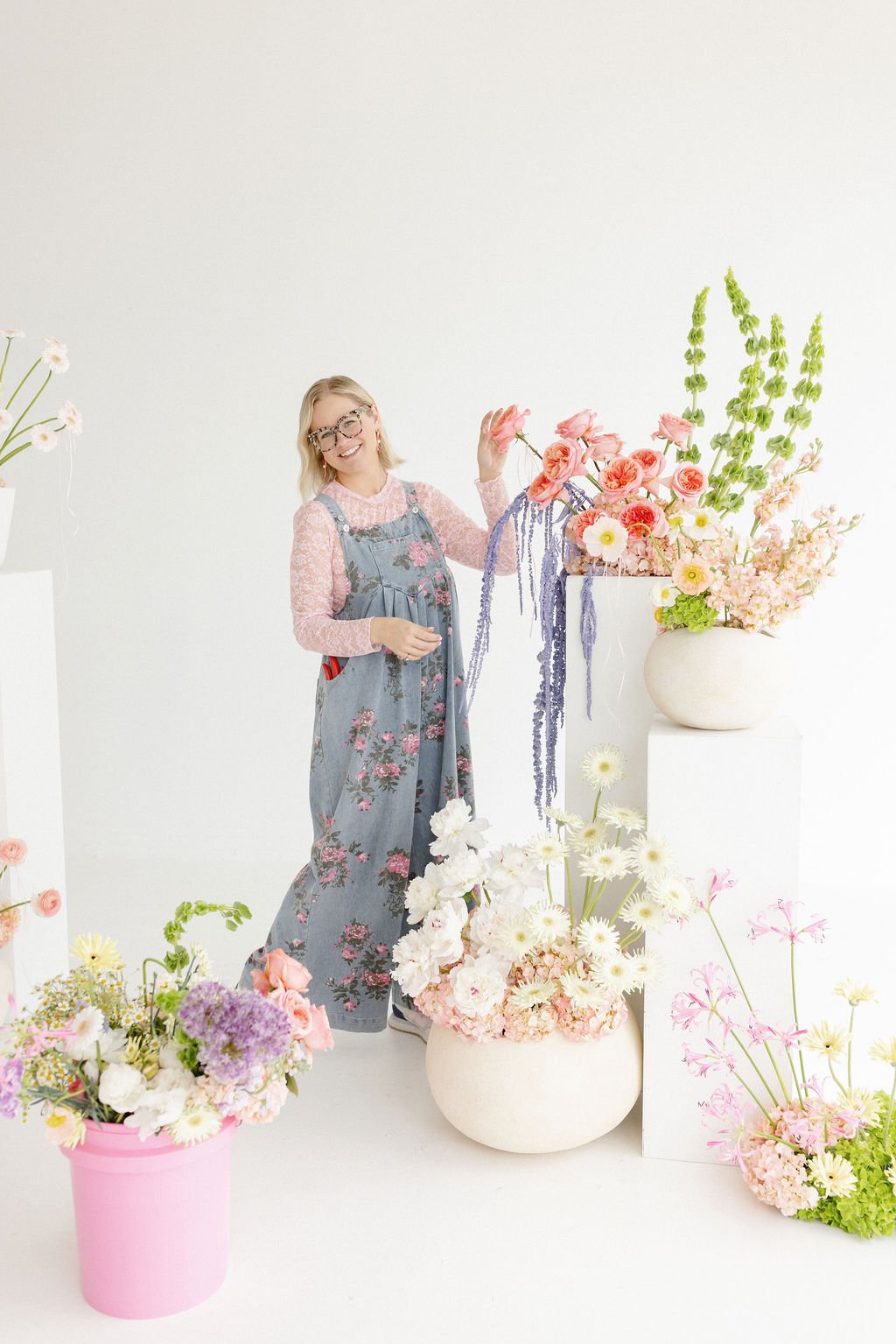 A woman with glasses wearing a pink long-sleeve shirt and denim overalls with floral pattern stands next to large flower arrangements in white vases, smiling and smiling in a bright, minimal studio setting.