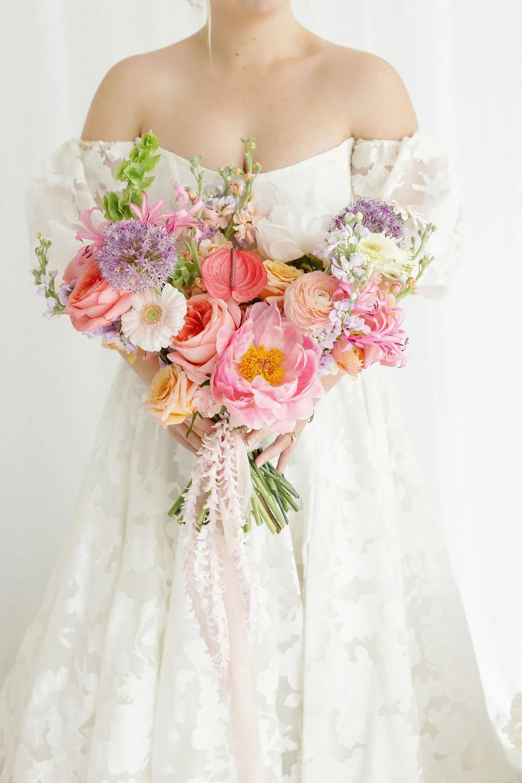 A woman in a white off-shoulder dress holds a bouquet of pink, purple, white, and peach flowers arranged in a heart shape, with some greenery.