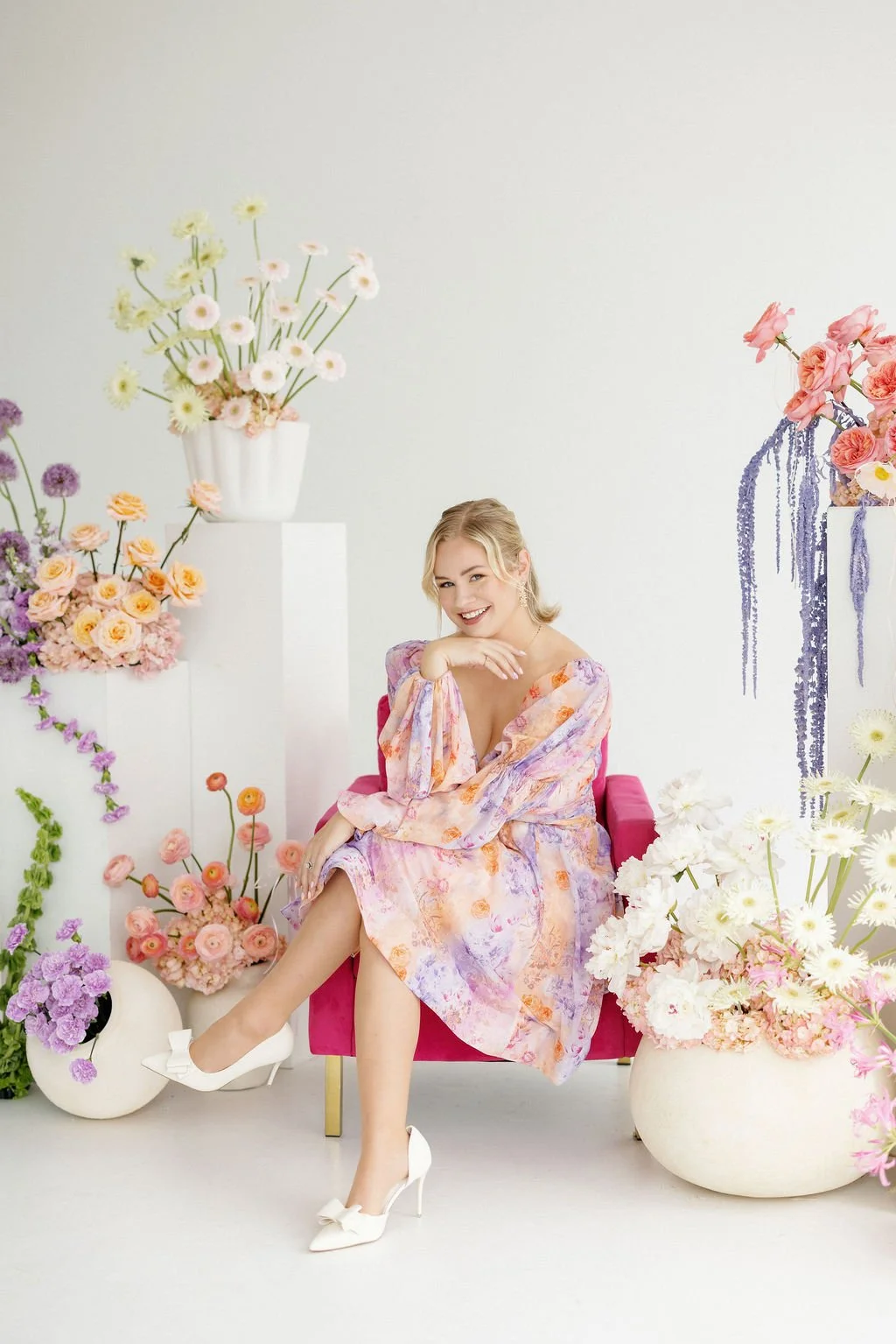 A woman sitting on a pink chair surrounded by colorful flowers in vases, smiling and posing in a bright, minimalist setting.
